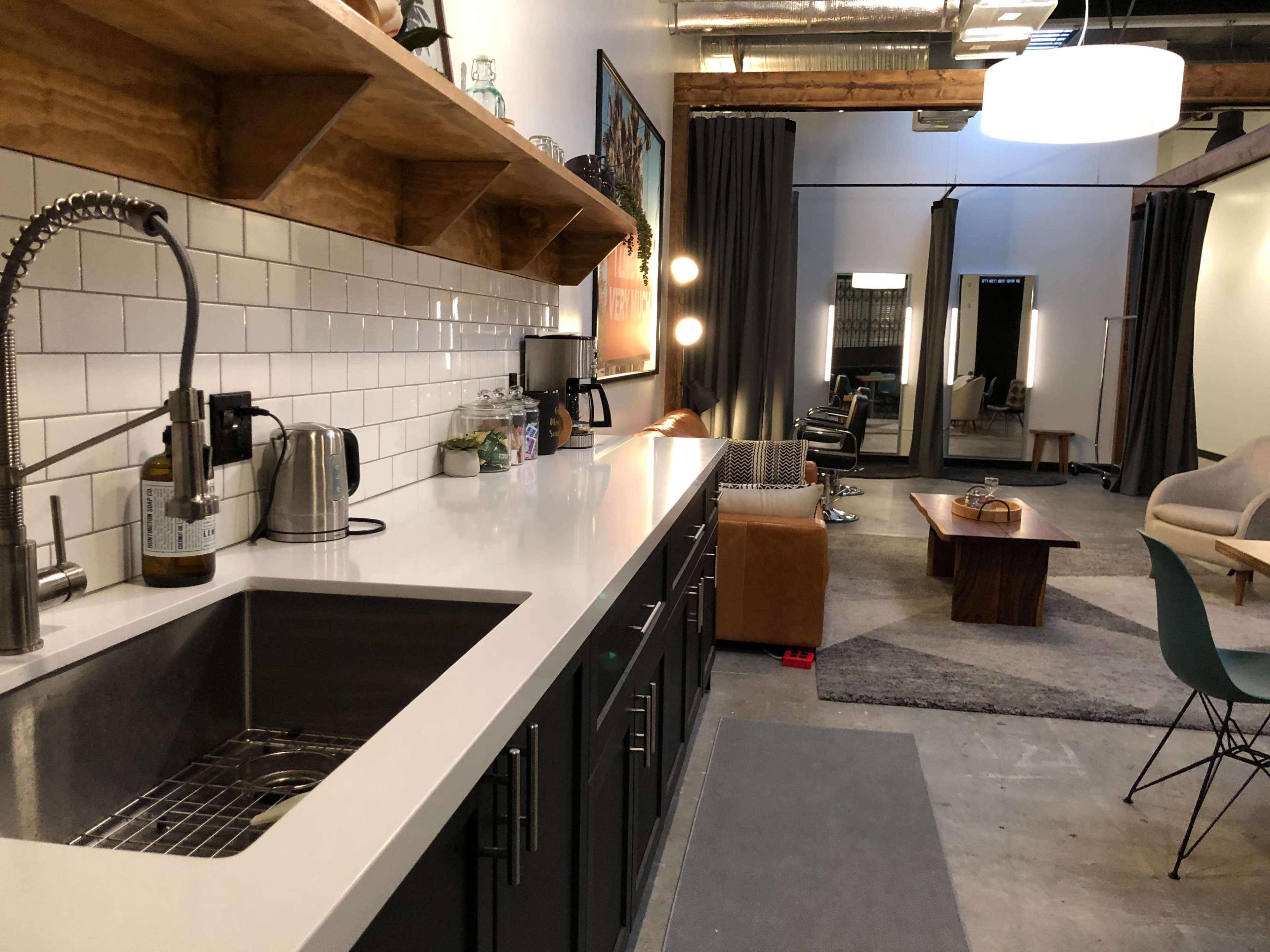 The image shows a modern kitchen with a dark cabinet and countertop, a stainless steel sink, and an open space leading to a living area with furniture and lighting fixtures.