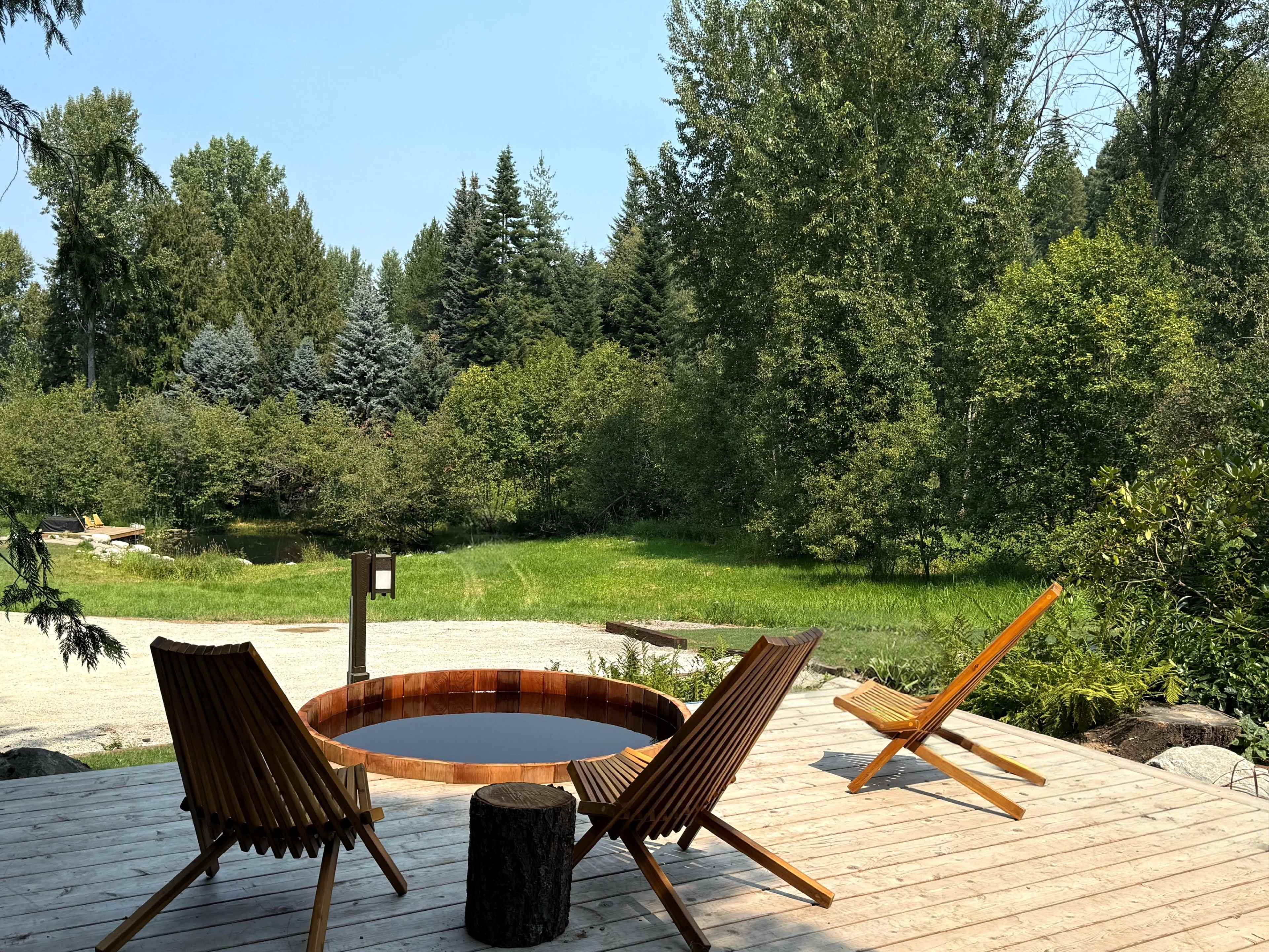 A wooden deck features a circular hot tub and two outdoor chairs overlooking a green landscape with trees.