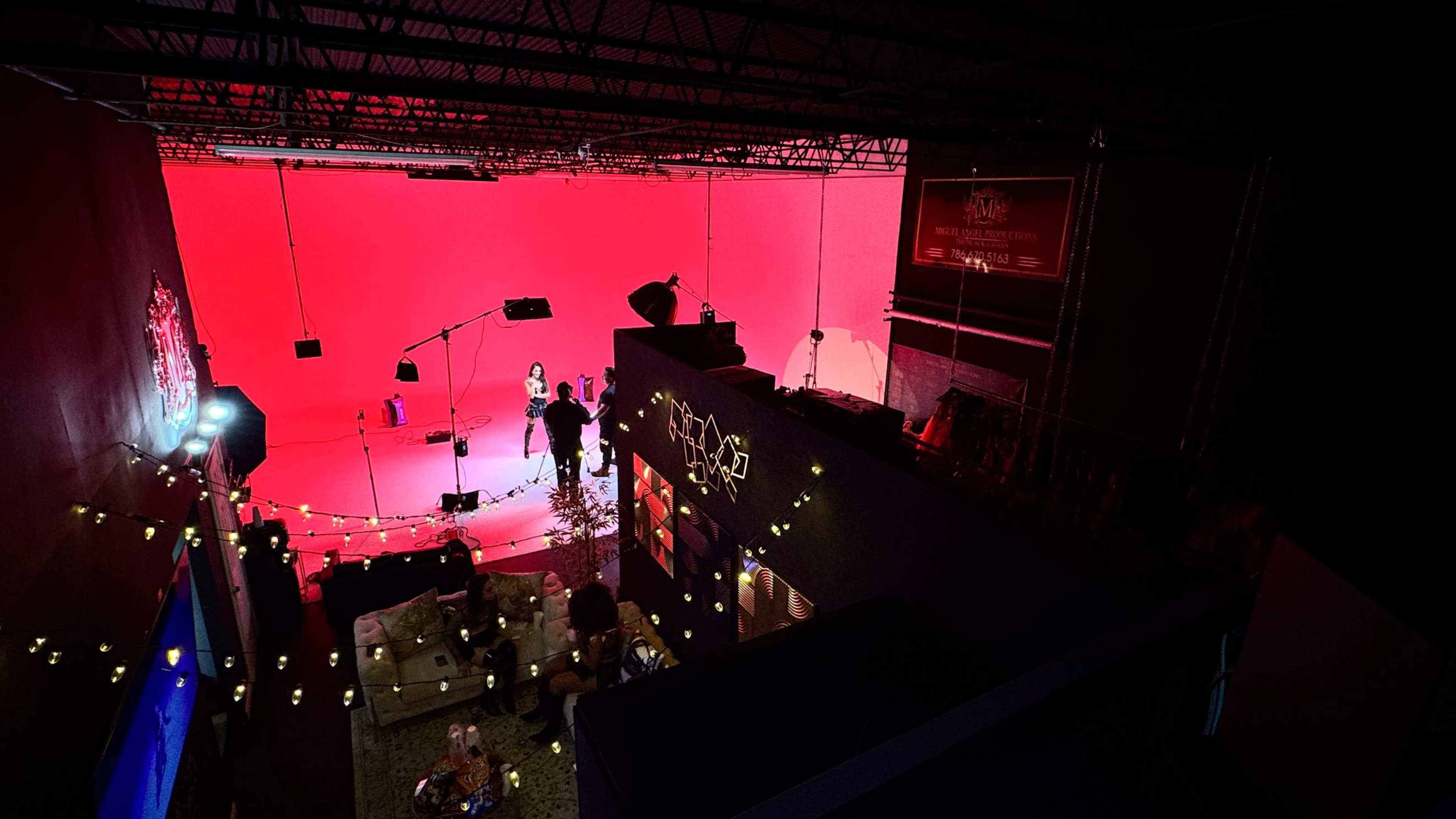 A brightly lit studio features a red backdrop, with equipment and crew preparing for a shoot while a lounge area is visible below.