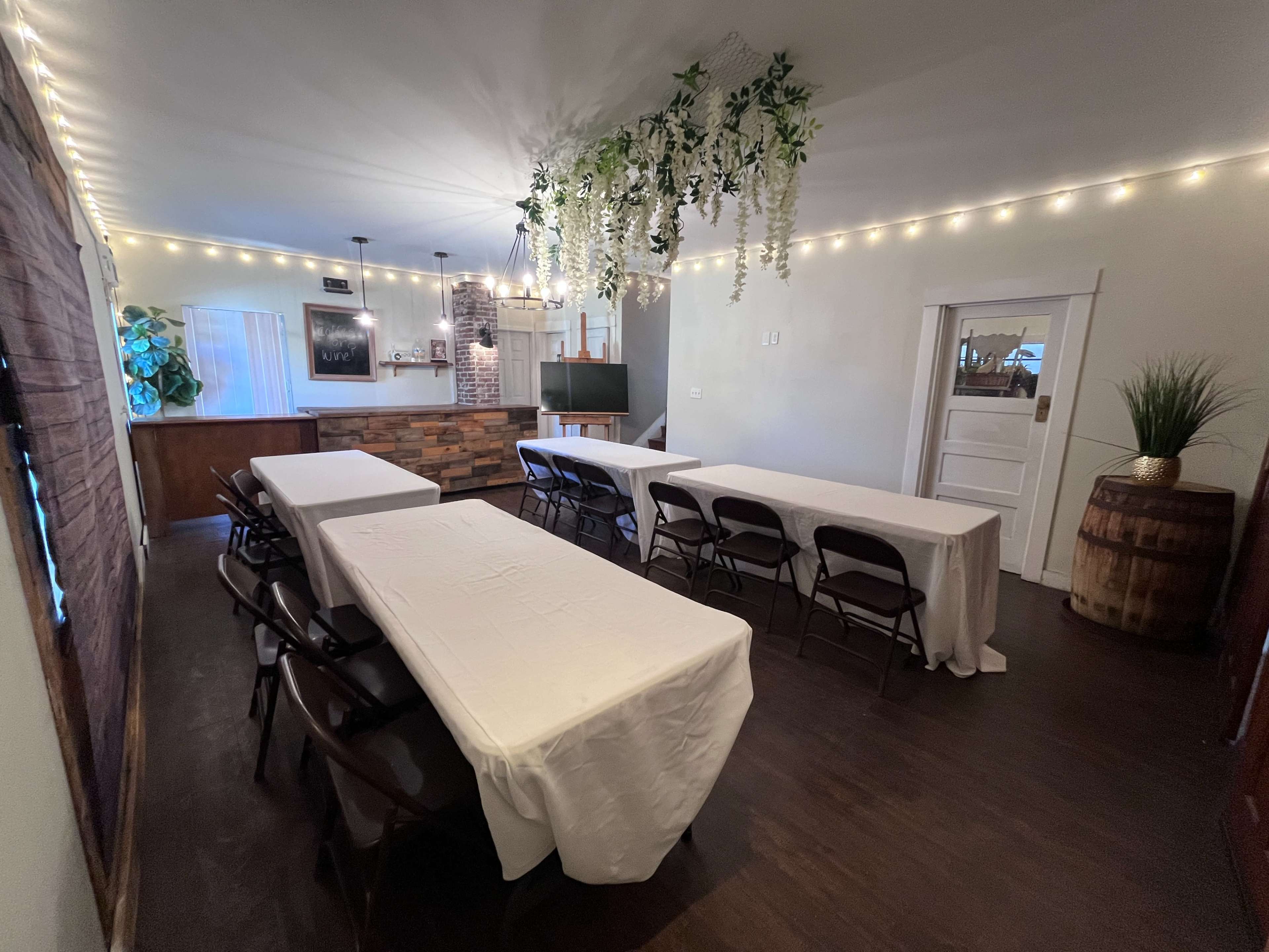 The image shows a room set up for an event, featuring several tables covered with white tablecloths and string lights hanging from the ceiling.