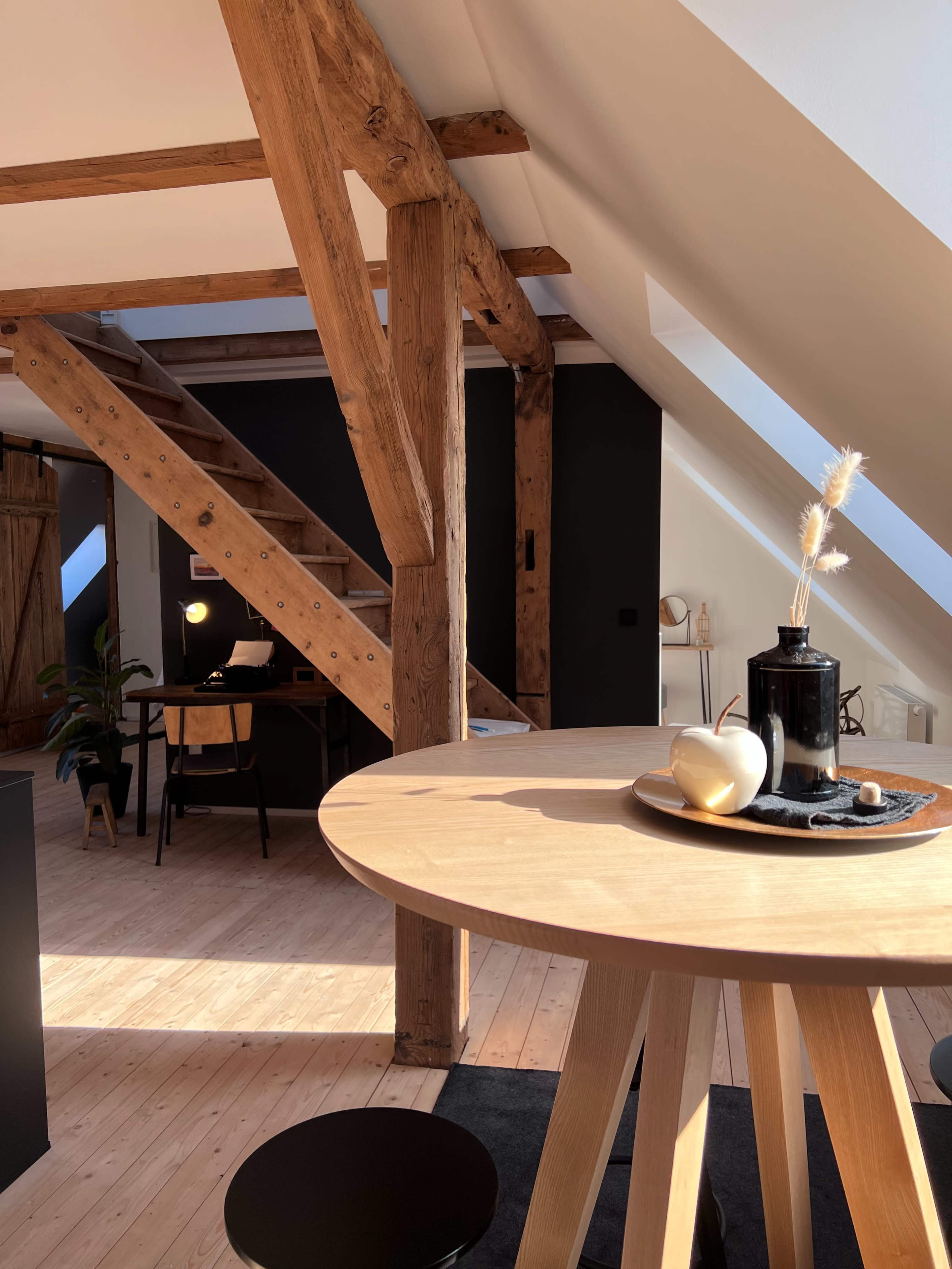 The image shows an interior designed space with wooden beams, a round table set with decorative items, and a staircase leading to a loft area.