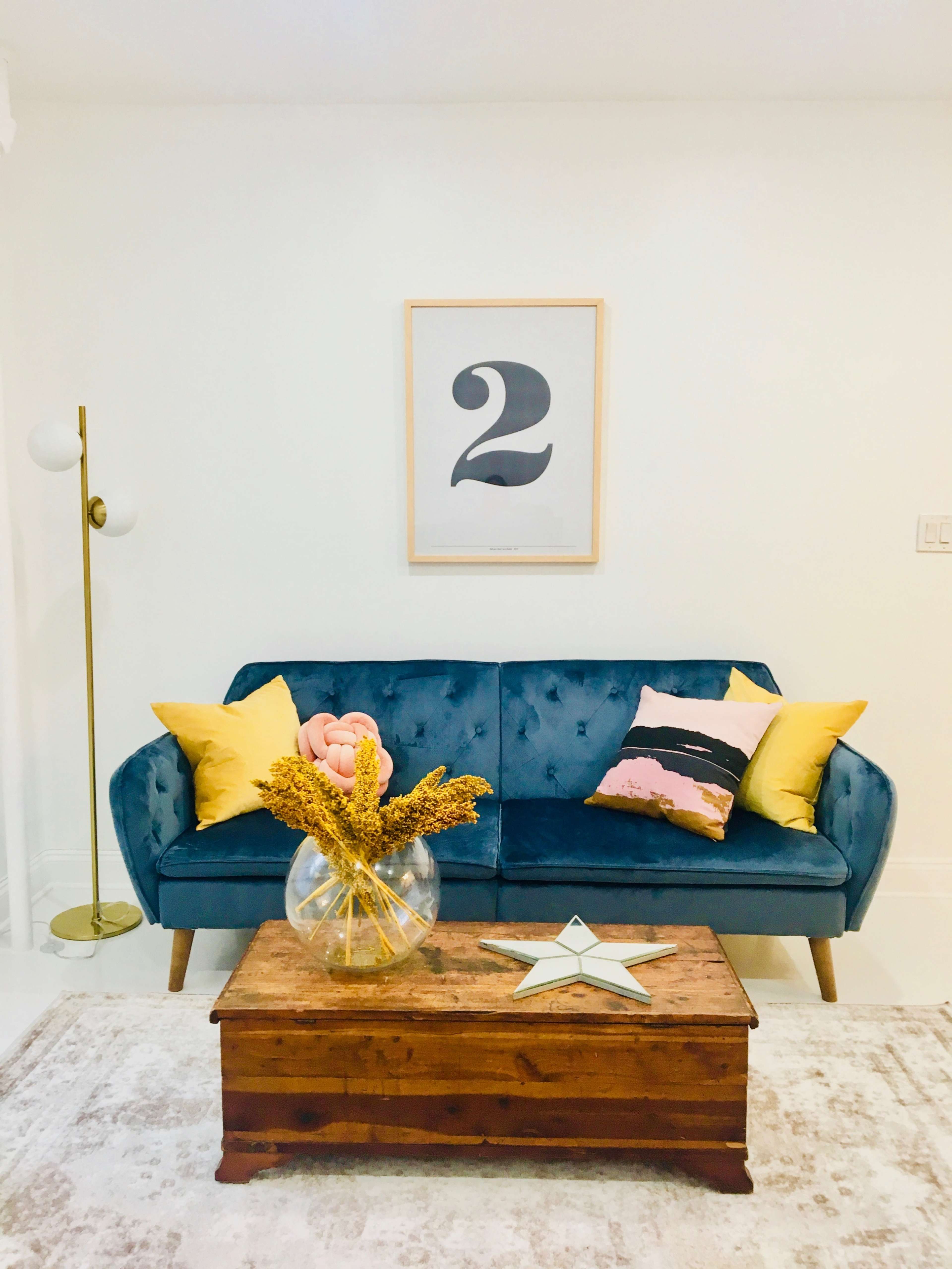 A blue velvet sofa with yellow pillows sits in a bright room next to a tall lamp and a wooden coffee table adorned with a vase of flowers and a decorative star.