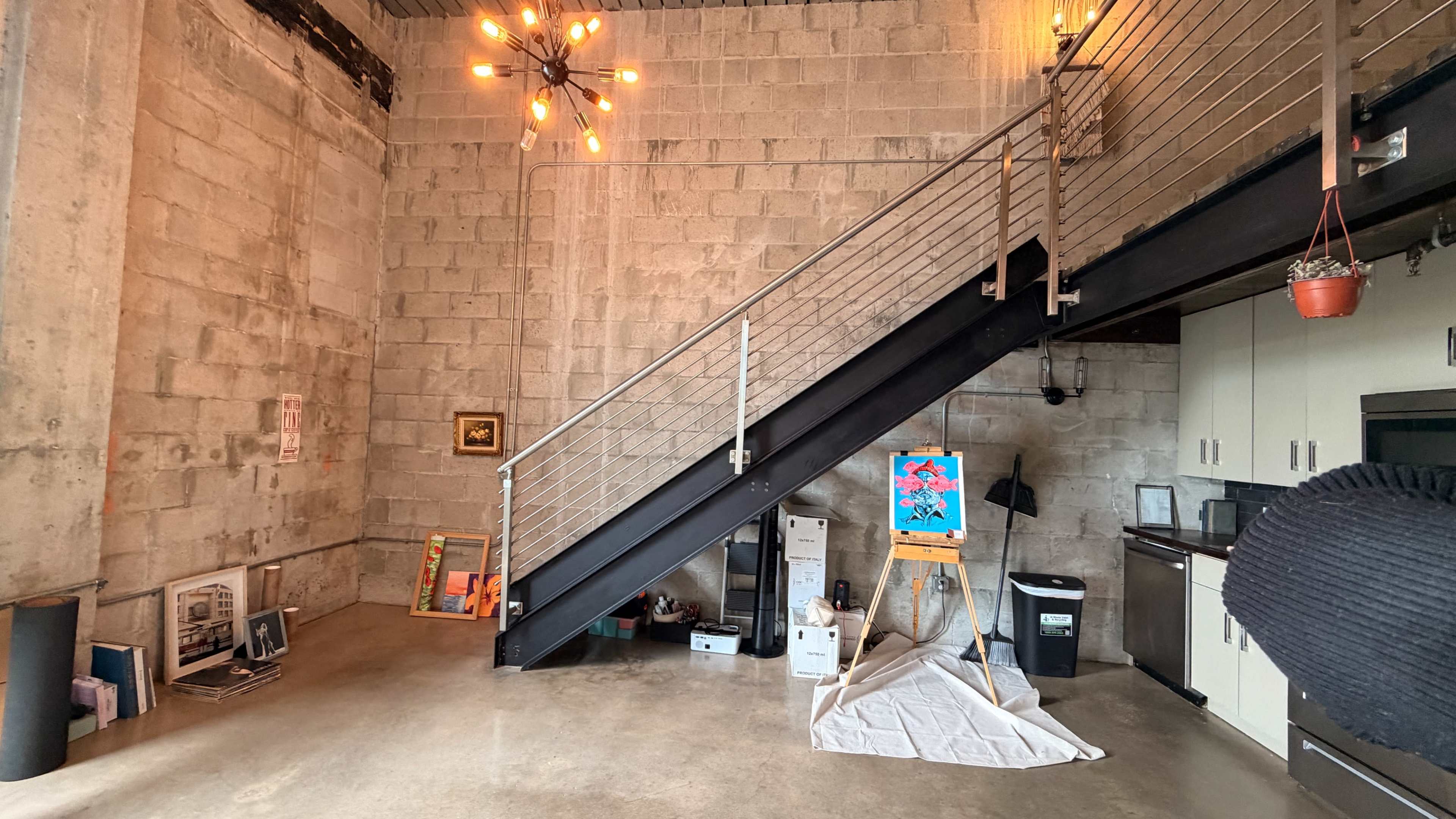 The image shows a modern interior space with a staircase leading to an upper level, featuring exposed concrete walls and various art supplies and artworks placed on the ground.