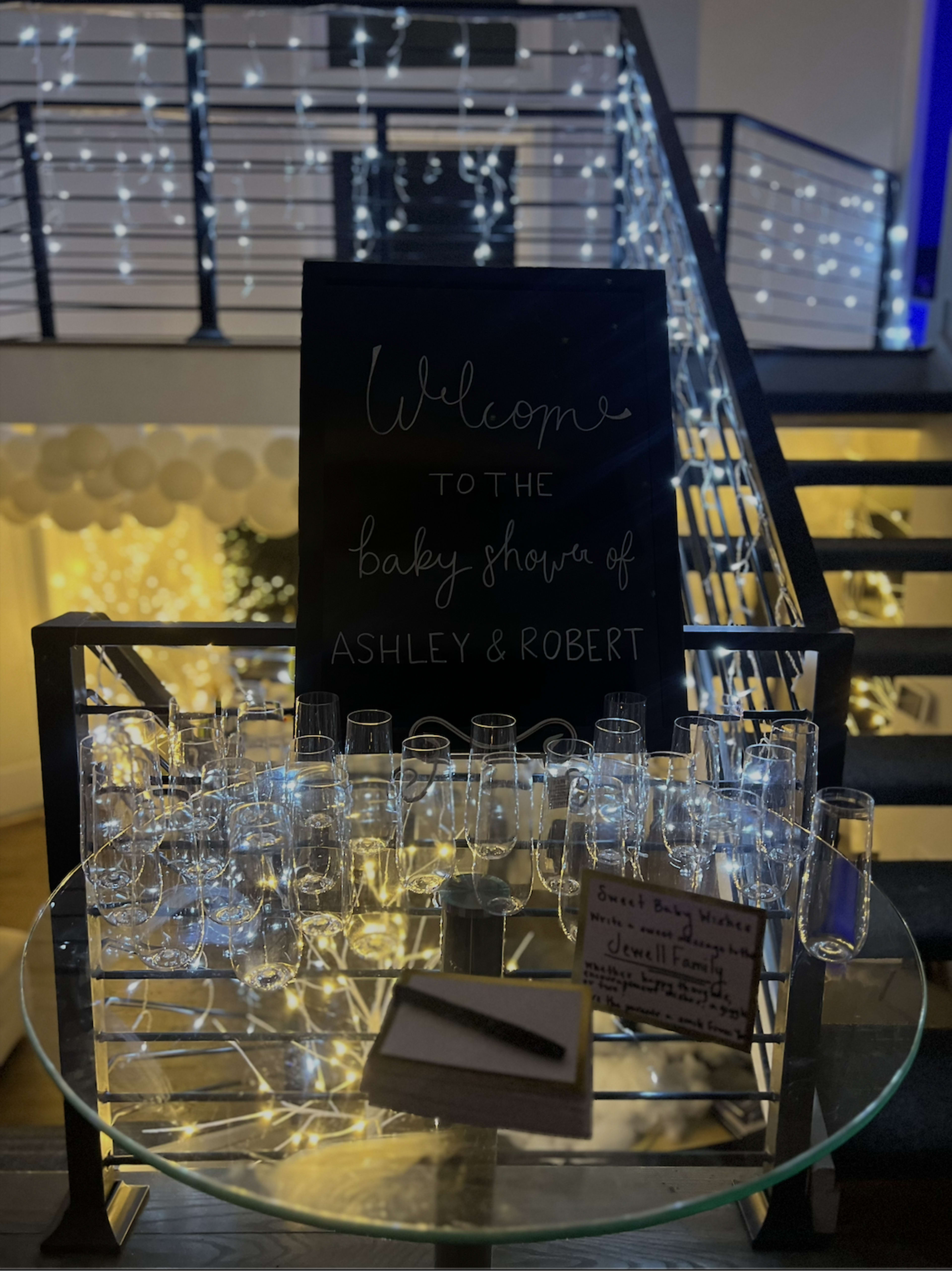 A welcome sign for a baby shower is displayed on a stand next to a collection of champagne flutes, with a staircase decorated with twinkling lights in the background.