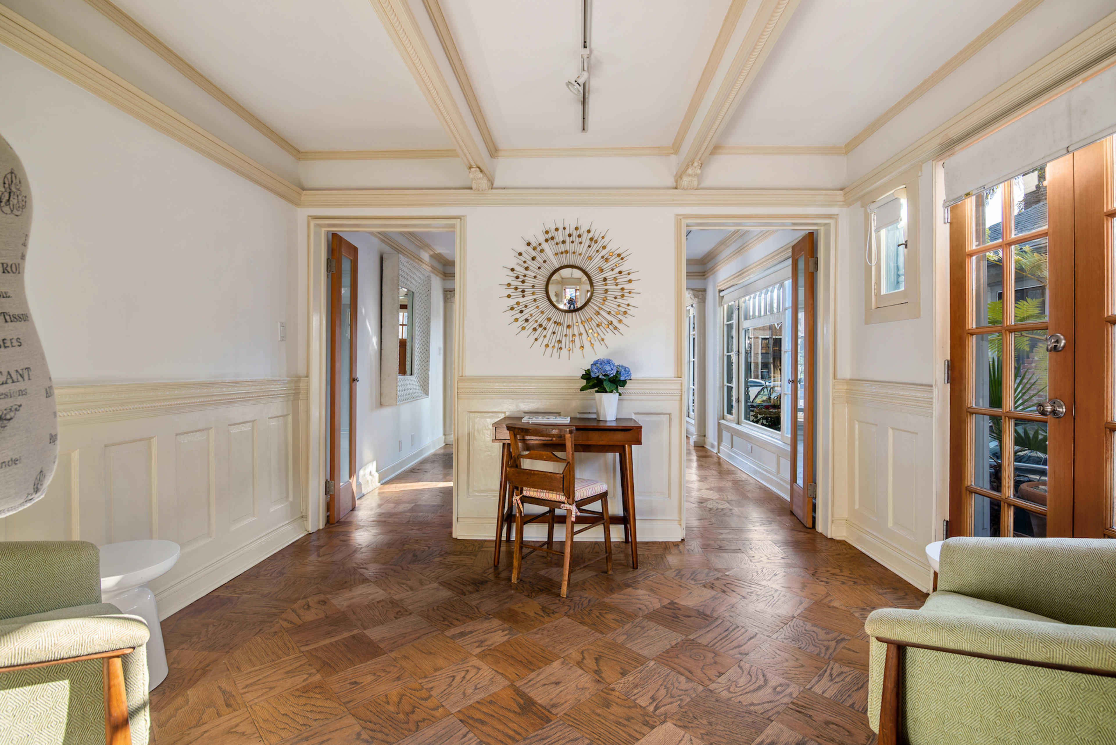 A well-lit hallway featuring a central table with a flower vase, flanked by two doorways, and wooden chairs on either side.