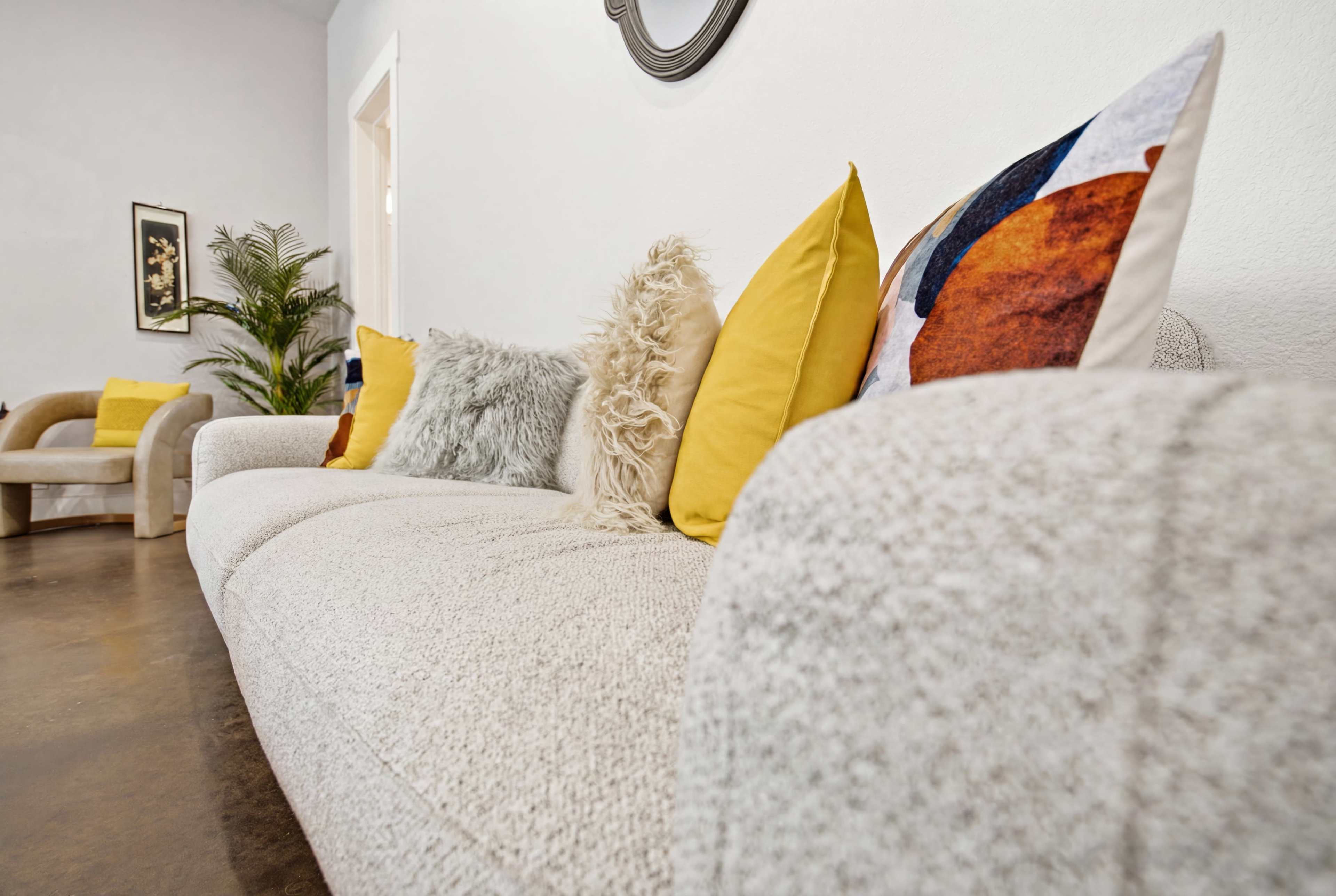 The image shows a modern living room with a textured white sofa adorned with colorful pillows, alongside a decorative plant and a framed artwork on the wall.