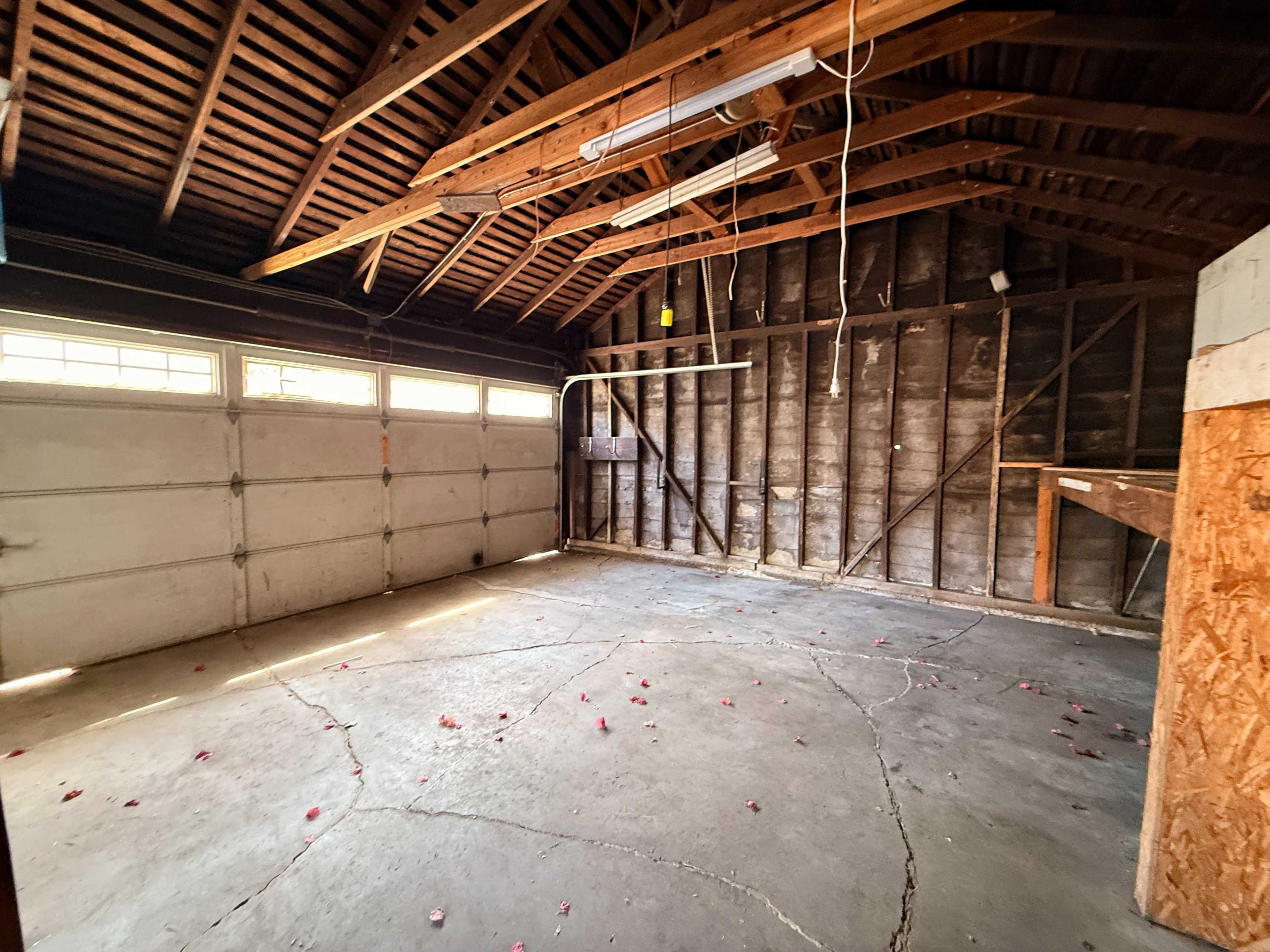 The image shows a spacious, empty garage with wooden beams and a concrete floor, featuring an overhead door and remnants of flower petals scattered across the ground.