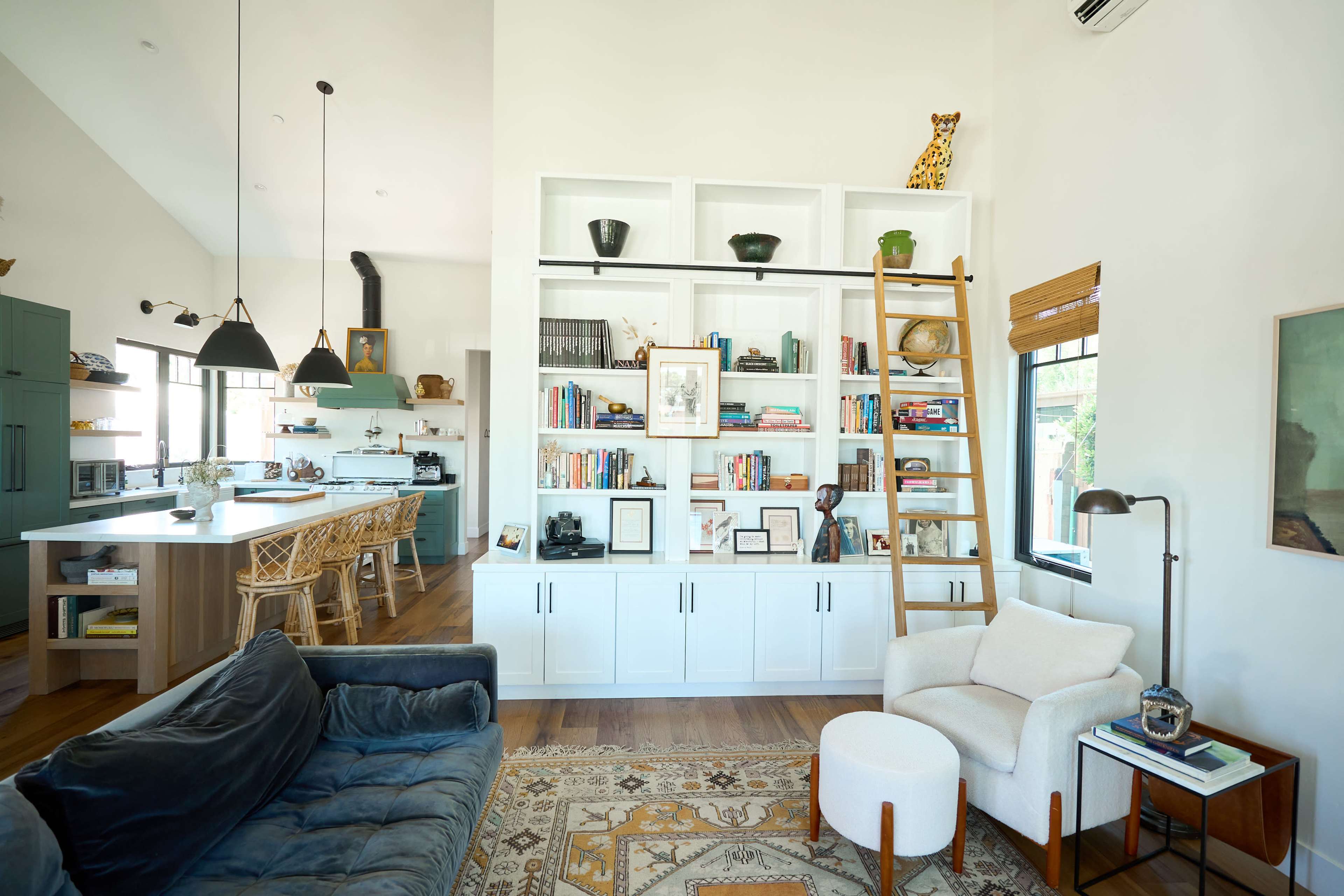 A cozy living space features a bookshelf filled with books and decor, a blue sofa, a white armchair, and a kitchen area in the background.