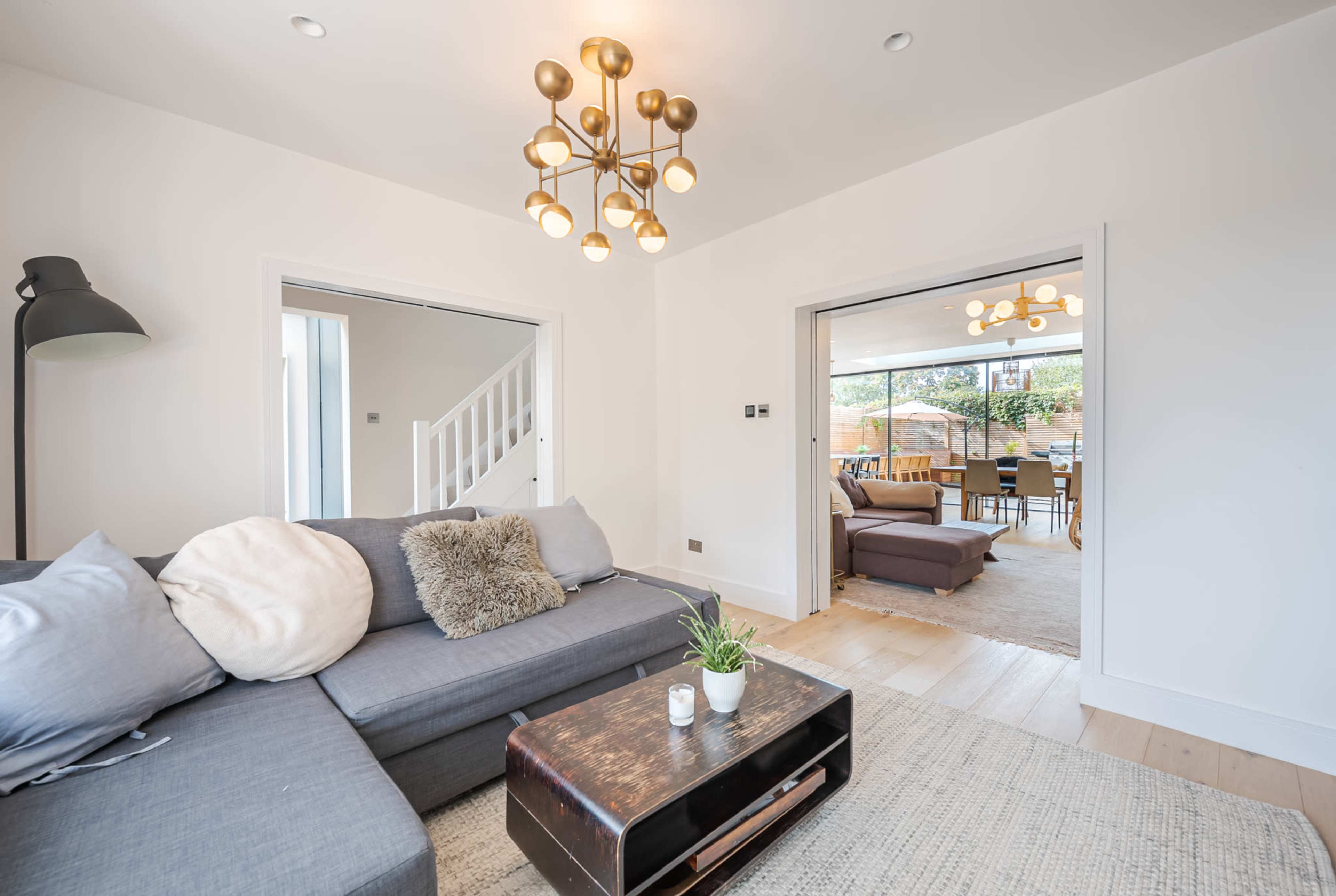 The image shows a modern living room with a sectional sofa, a coffee table, and a chandelier, leading to an open dining area visible through two doorways.
