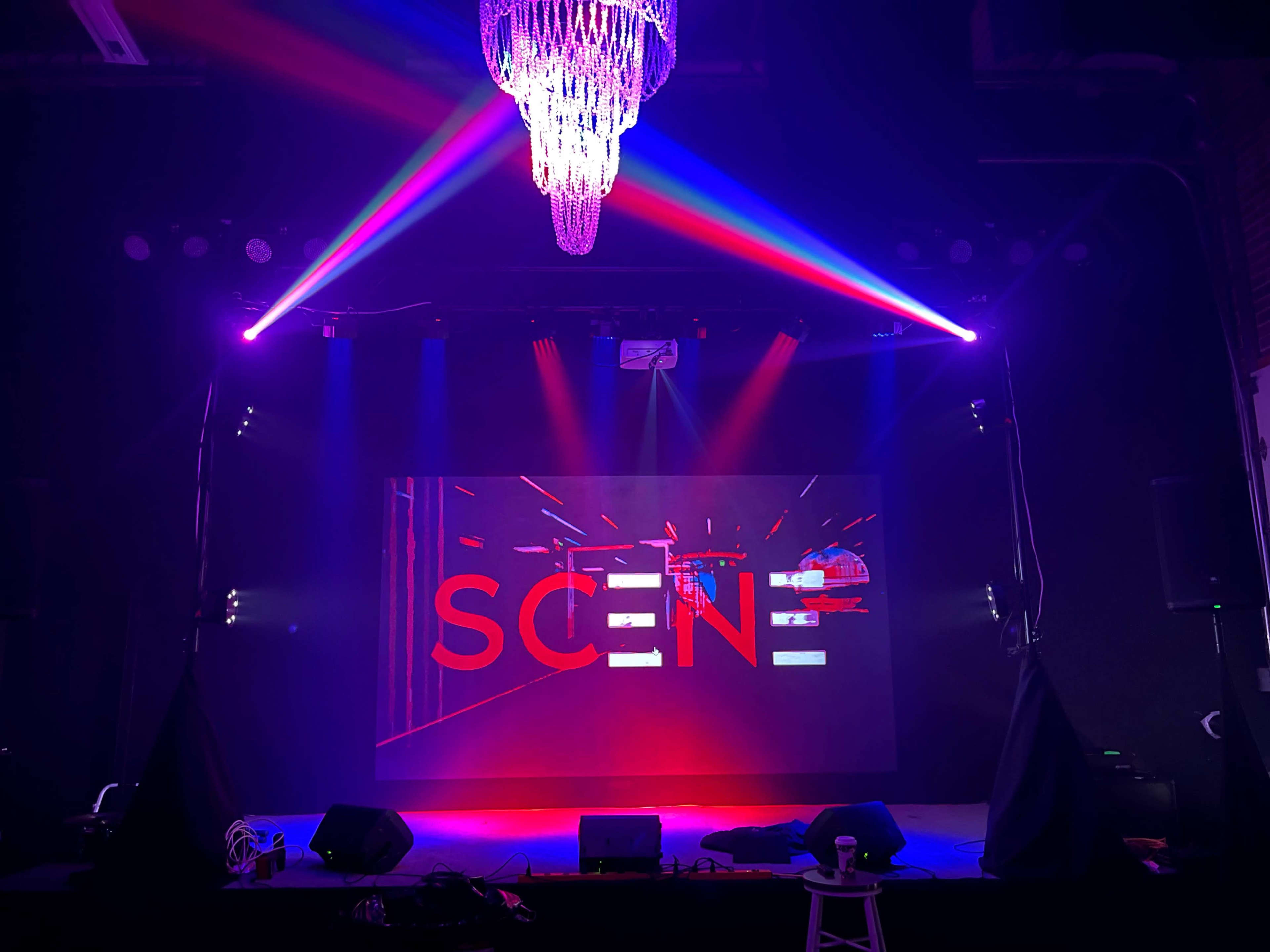 A stage with a large screen displaying the word "SCENE," illuminated by colorful lights and featuring a chandelier overhead.