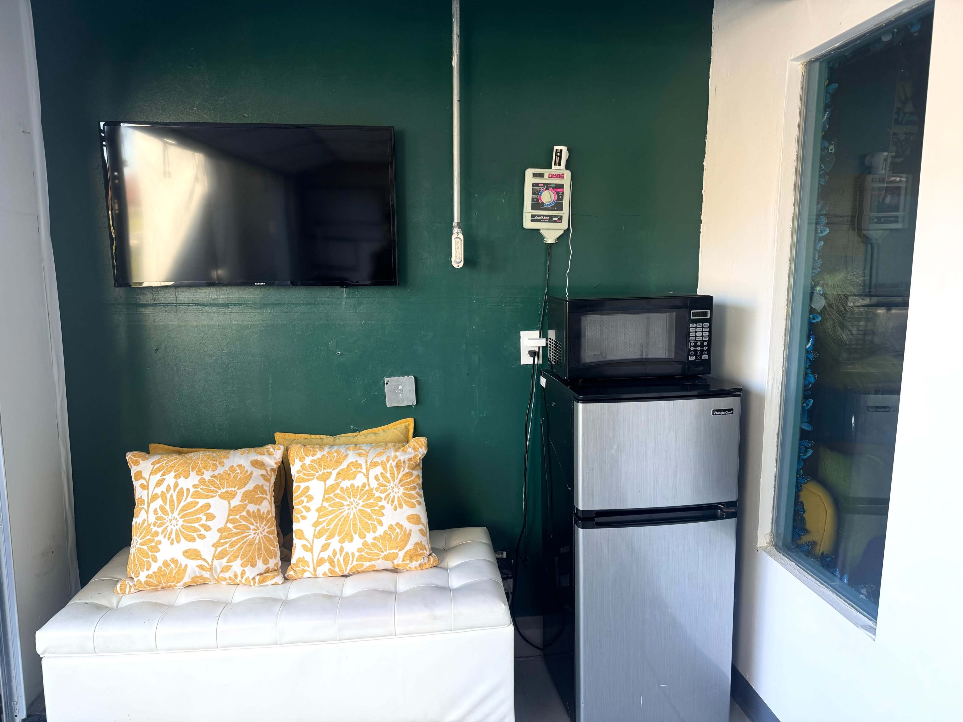 The image shows a small room with a green wall, a wall-mounted TV, a white bench with yellow floral cushions, a microwave, and a mini refrigerator.