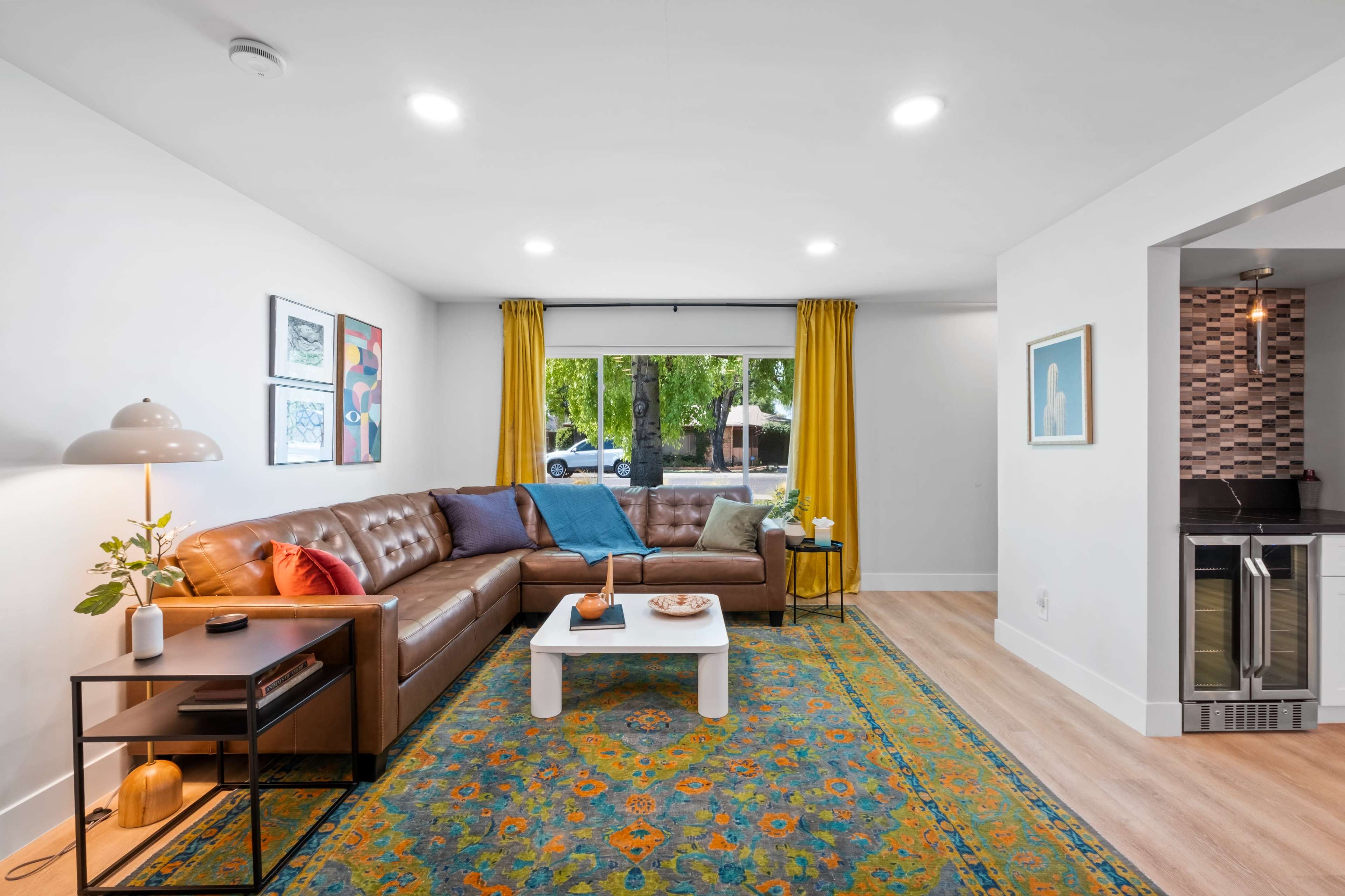 A modern living room featuring a brown leather sectional sofa, a white coffee table, and a patterned area rug, with yellow curtains framing a window that allows natural light to enter.