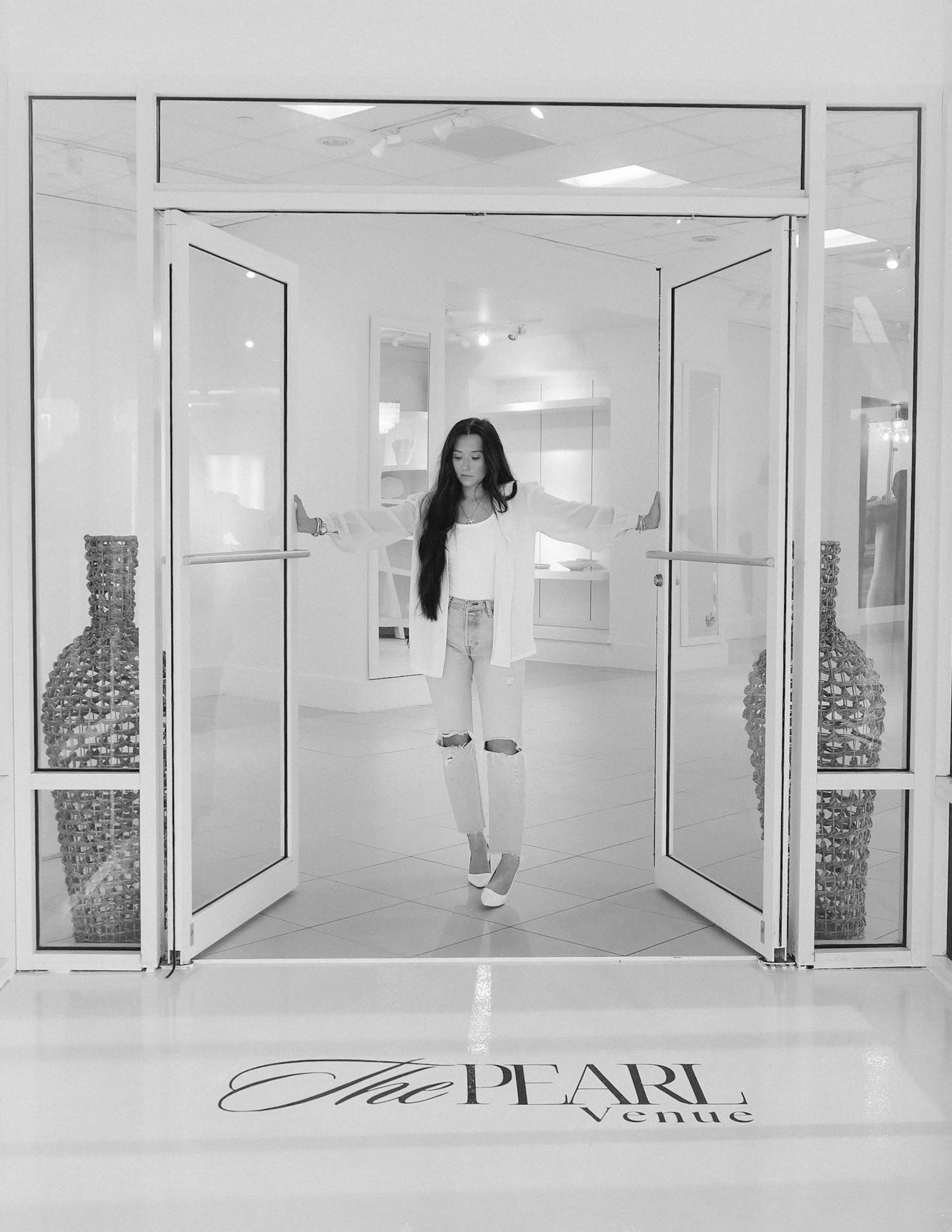 A woman stands in the doorway of a modern, white venue, framed by large glass doors and decorative vases on either side.