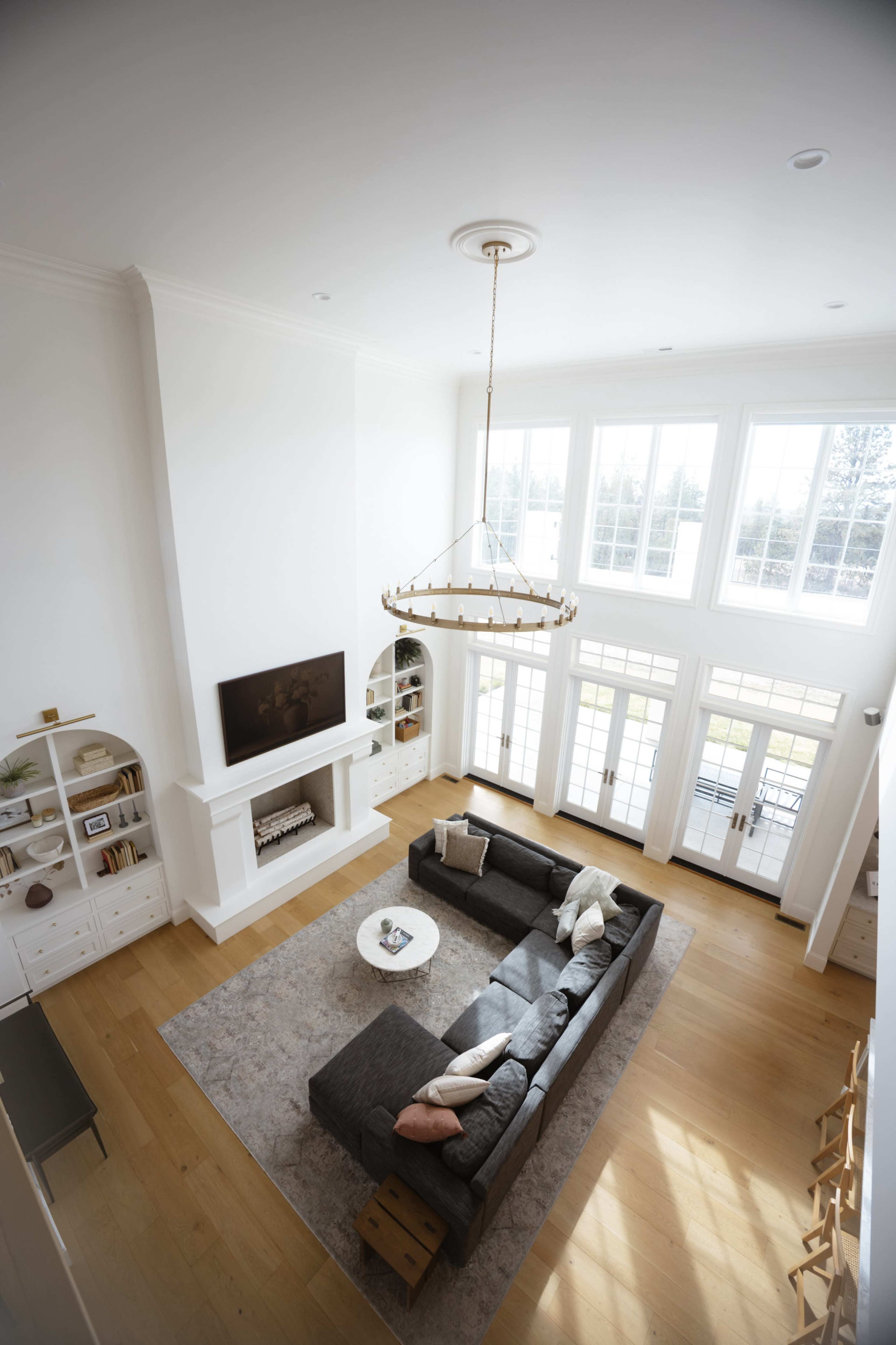 The image depicts a spacious living room with a high ceiling, a large sectional sofa, a coffee table, and multiple windows allowing natural light to fill the space.