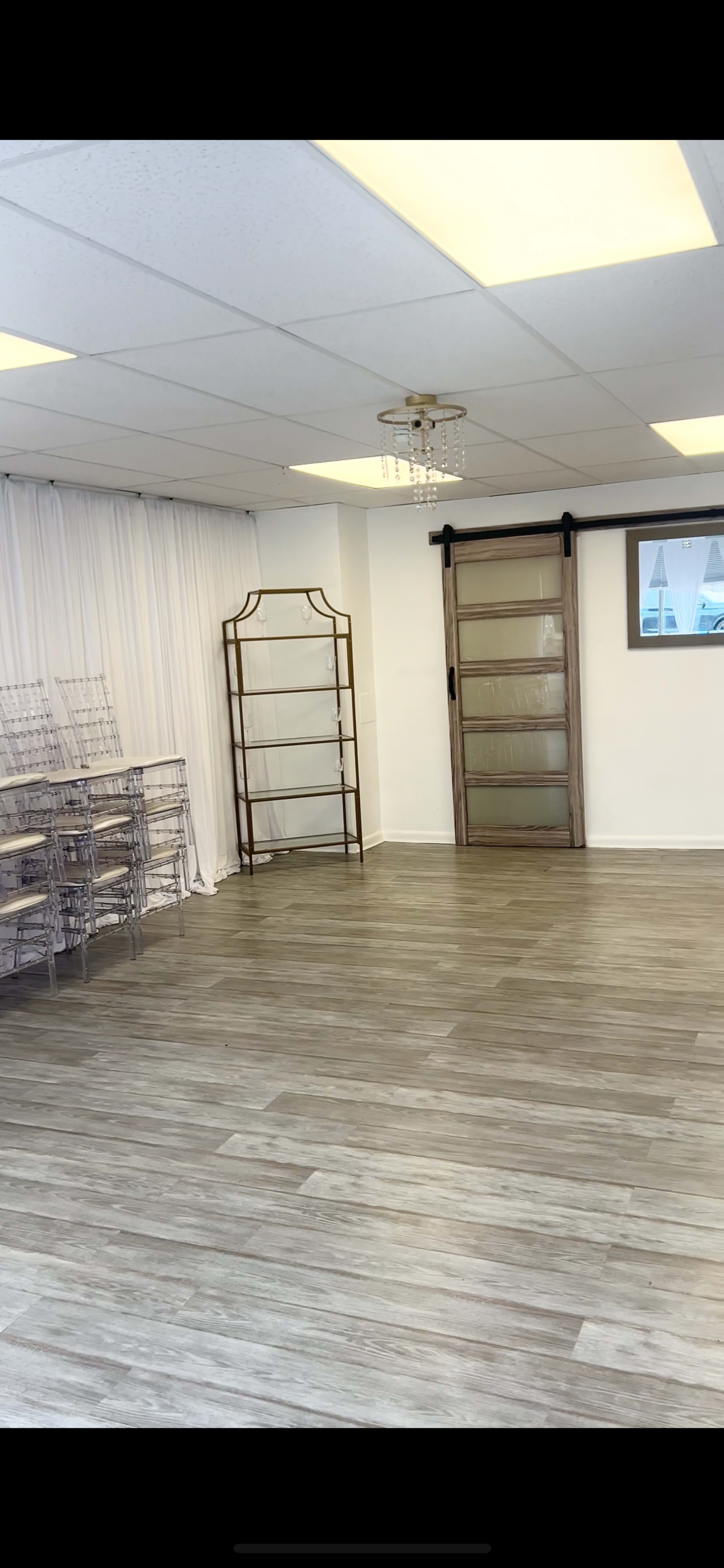 The image shows a spacious, empty room with light-colored wood flooring, a white draped wall, a metal shelving unit, and a sliding glass door.