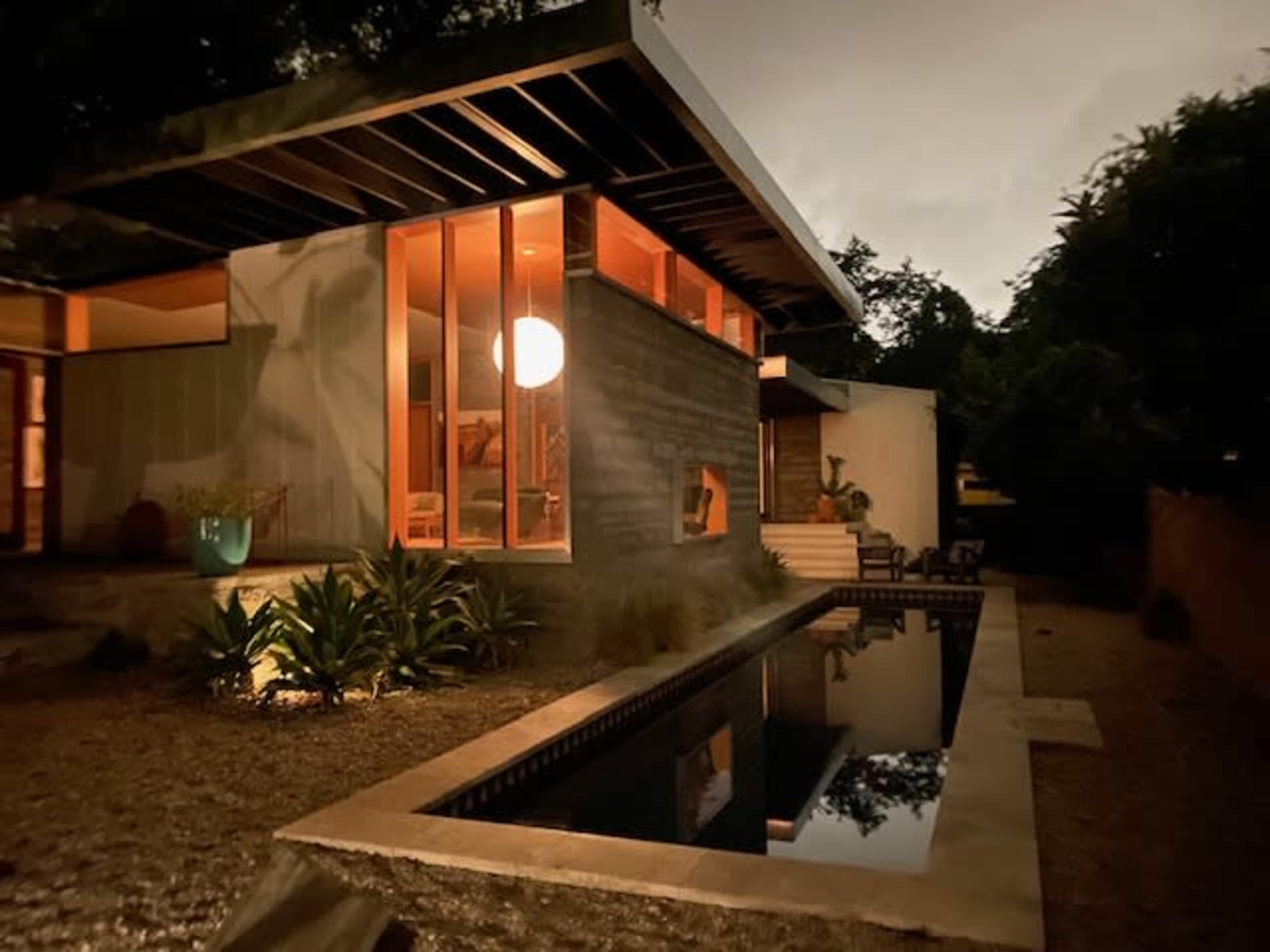 A modern house with large windows and a swimming pool reflecting the structure, surrounded by greenery in a dimly lit environment.