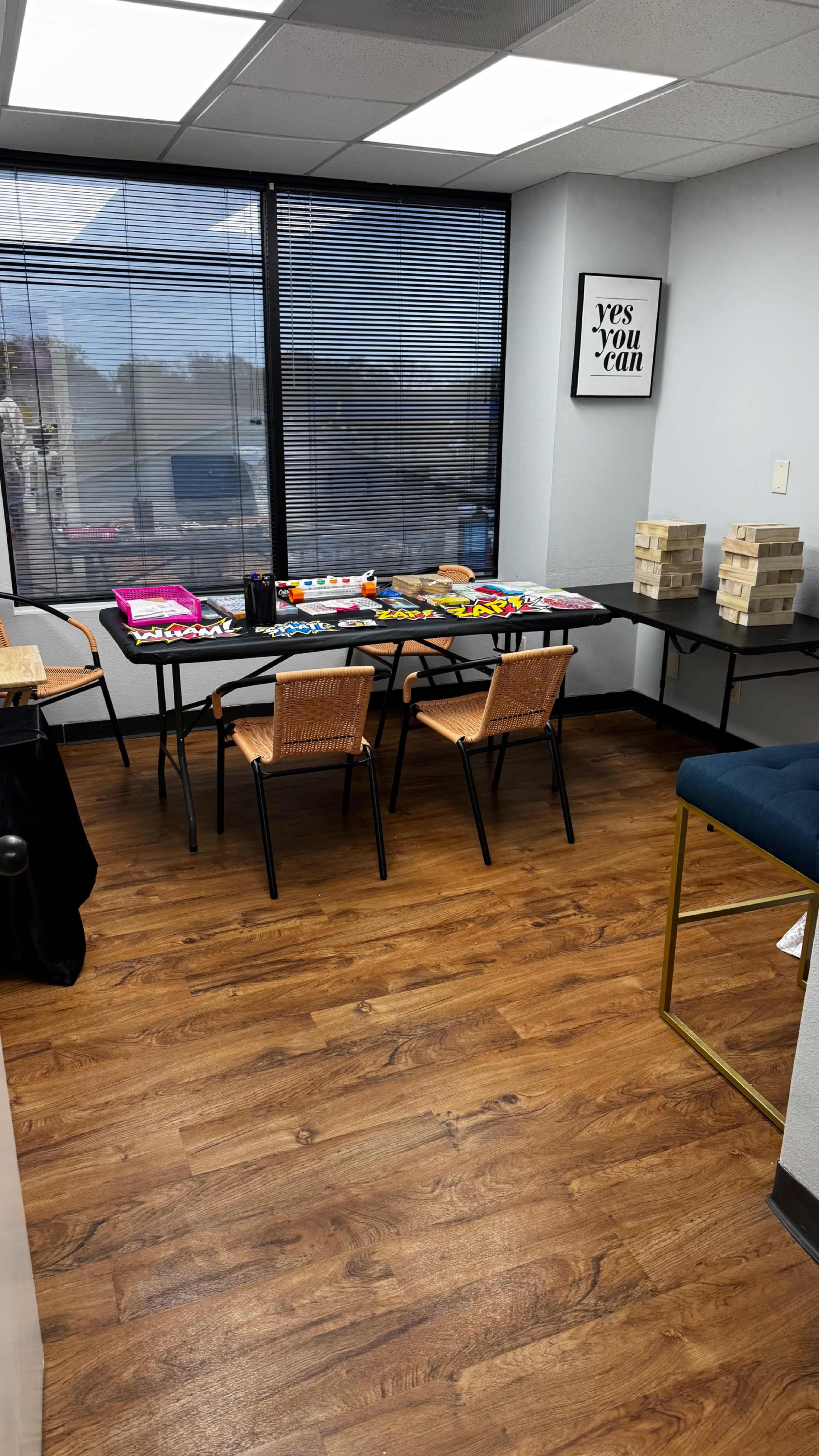 The image shows a small room with wooden flooring containing a table set with colorful games, two chairs, and a nearby stack of Jenga blocks.