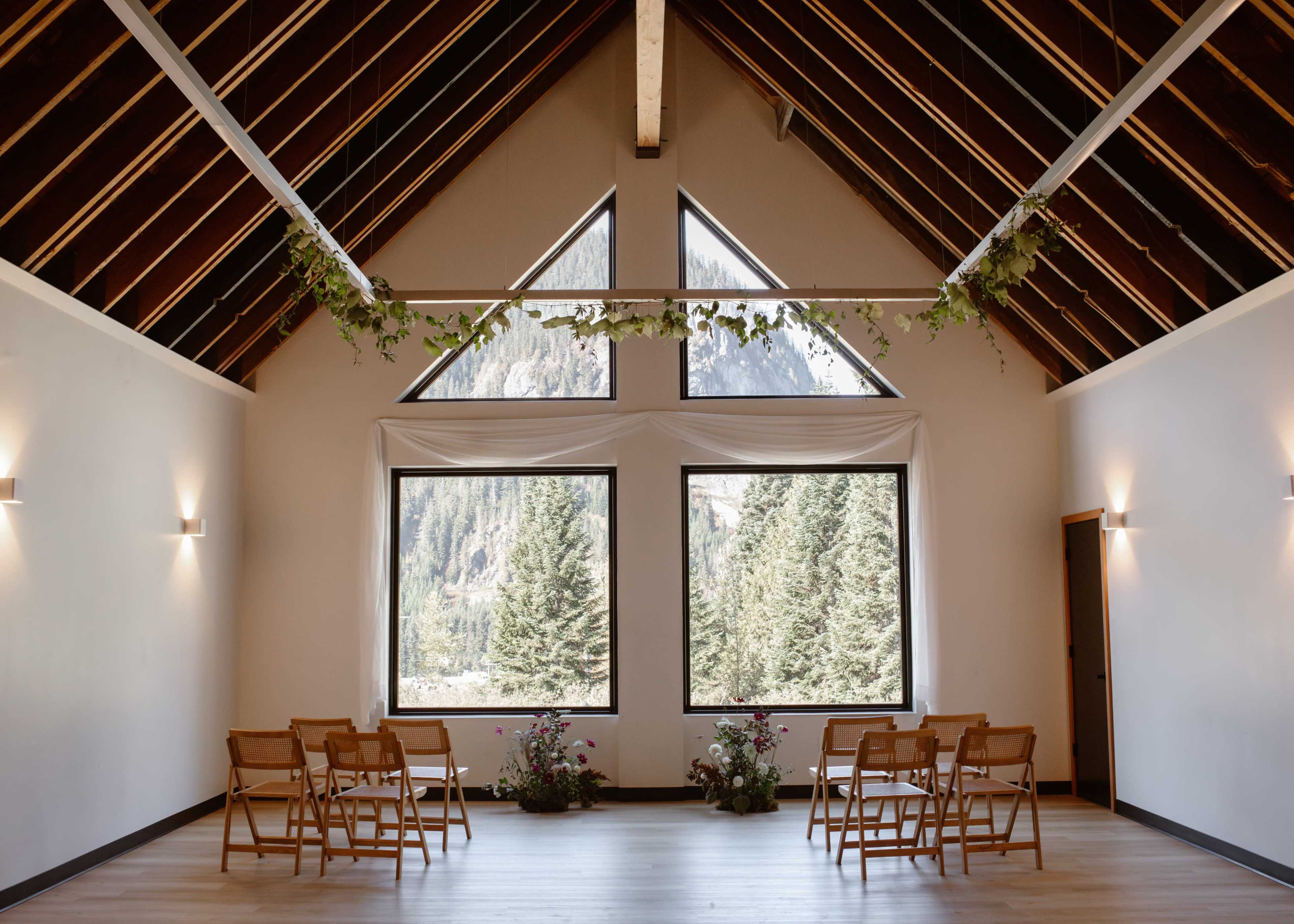 A spacious room with a vaulted ceiling features large windows overlooking trees and mountains, accompanied by wooden chairs arranged in front of floral decorations.