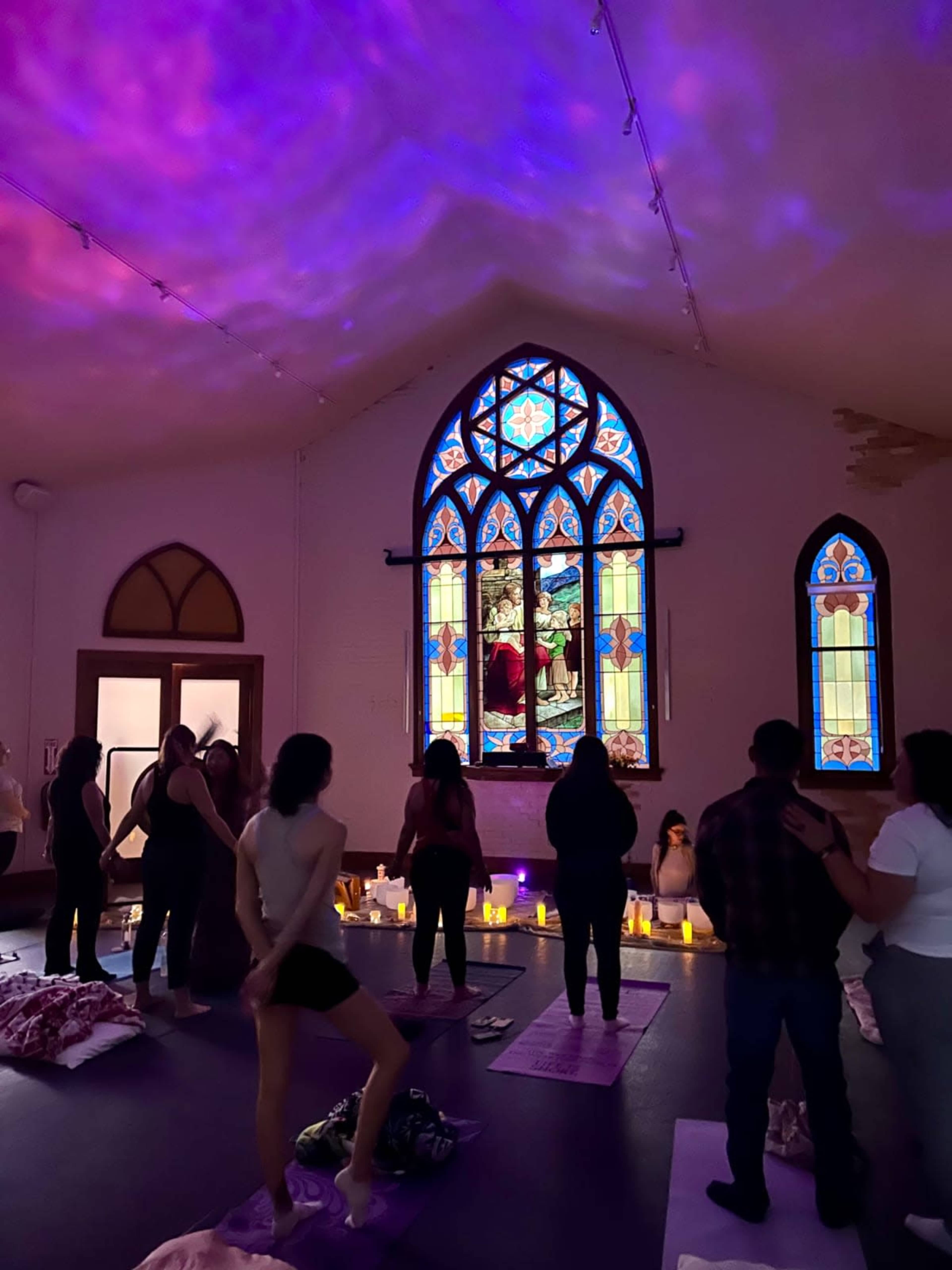 A group of people practice yoga in a dimly lit room featuring a large stained glass window and candles arranged on the floor.