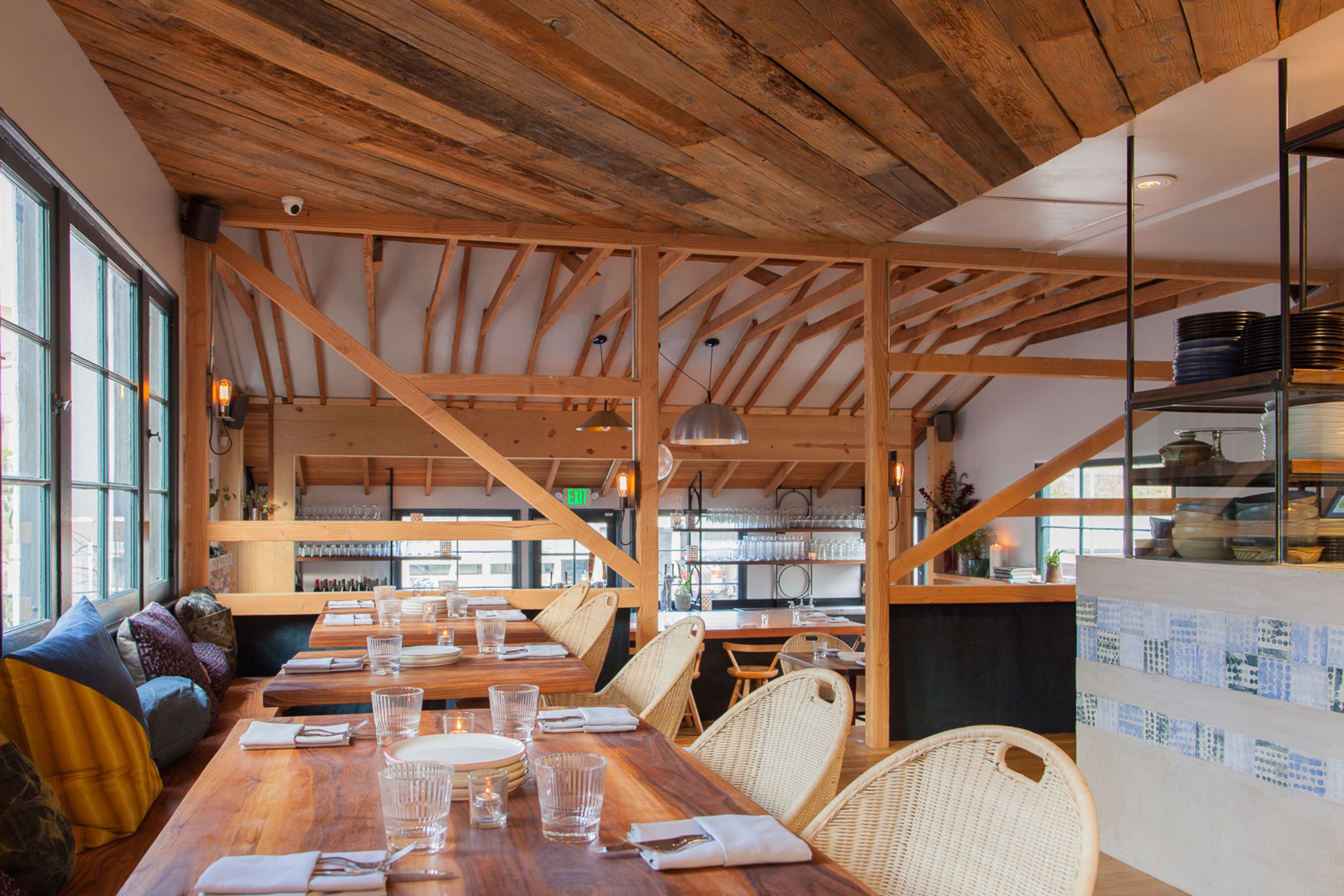 A spacious restaurant interior features wooden beams, dining tables set with plates and glasses, and a bright atmosphere enhanced by large windows.