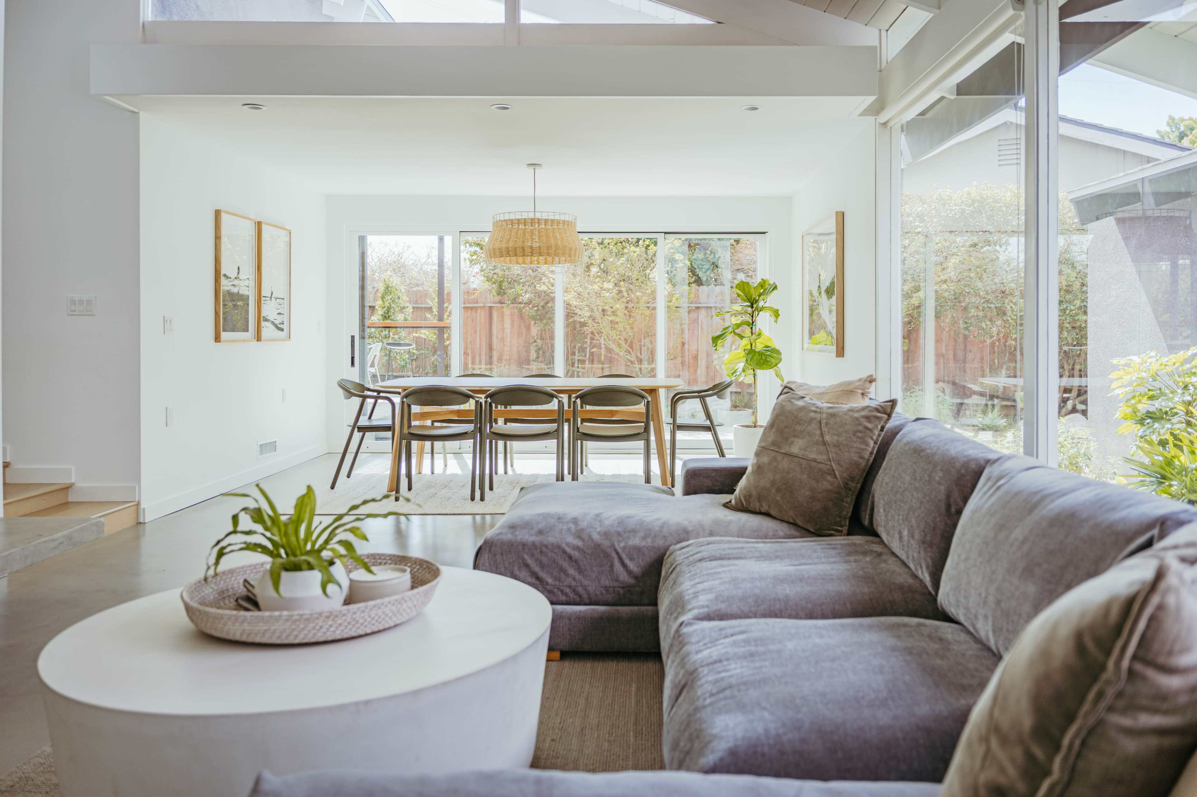 A modern living space features a gray sectional sofa, a round coffee table with a plant, and a dining area with a large table illuminated by a pendant lamp.
