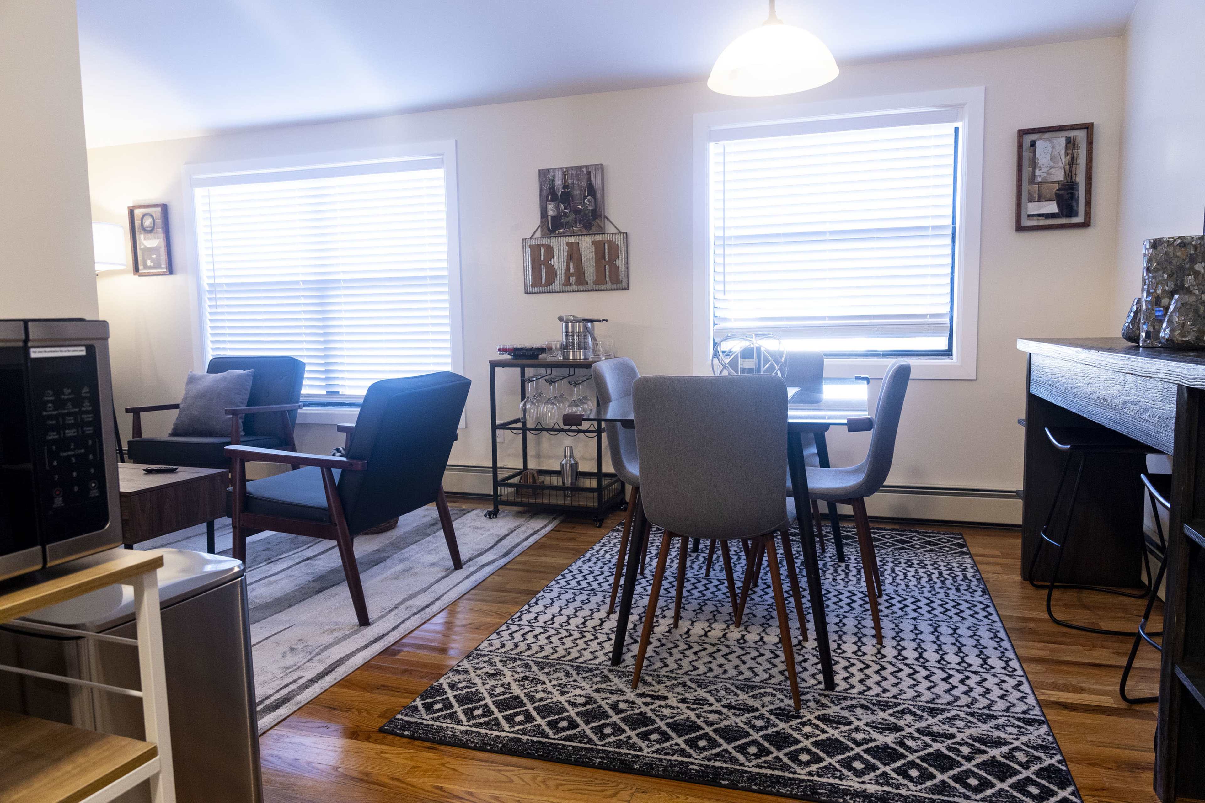 A modern living space featuring a seating area with two armchairs, a dining table with four chairs, and various decorative elements, all illuminated by natural light from two windows.