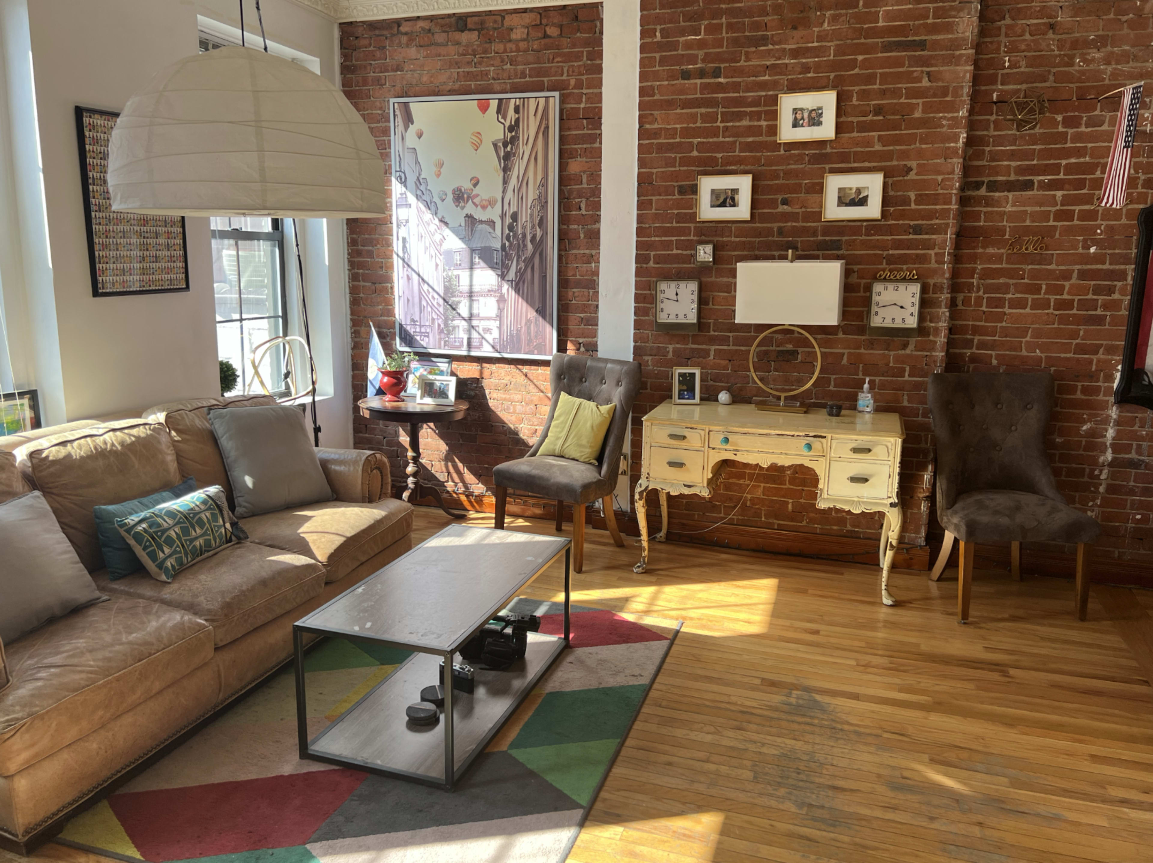 A living room with a beige sectional couch, a coffee table, two upholstered chairs, and a console table against a brick wall.