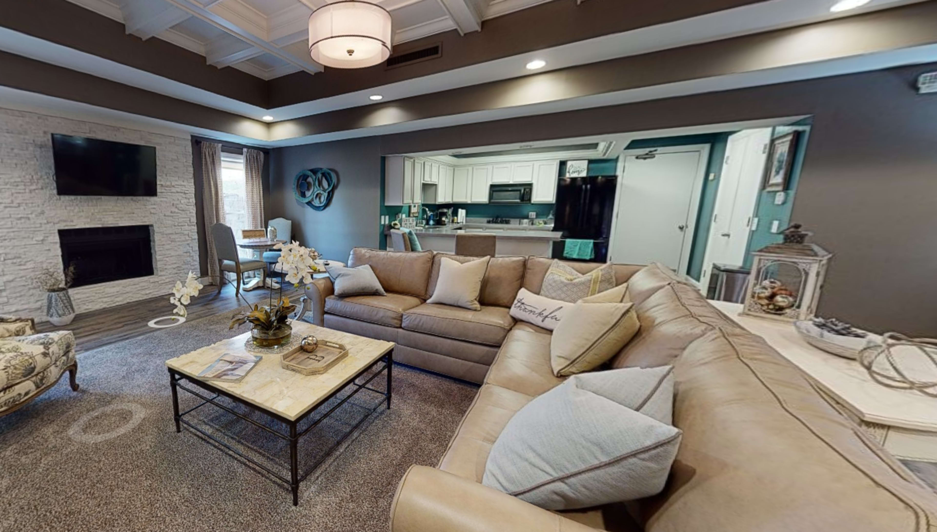 A living room with a beige sectional sofa, a coffee table, and a fireplace, leading into a kitchen area visible in the background.