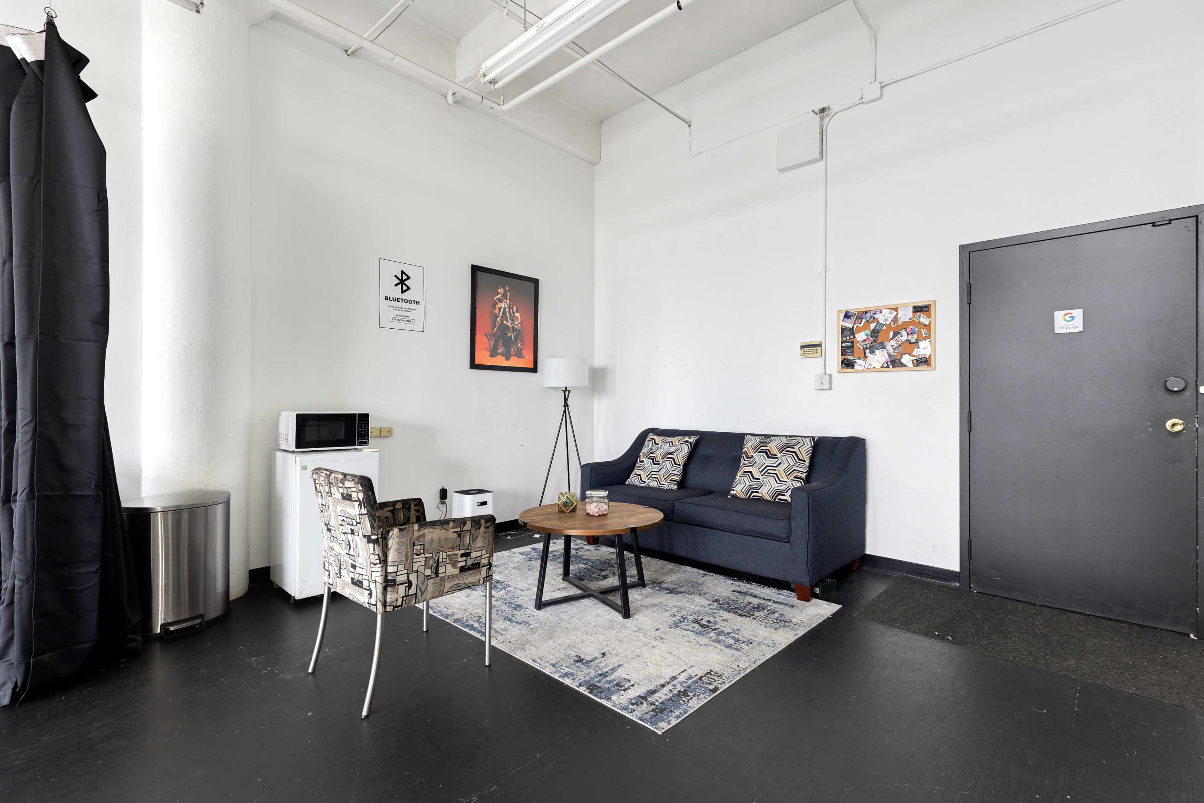 The image shows a small, minimalist lounge area with a dark sofa, a coffee table, a microwave, and a chair beside a door in a well-lit room.