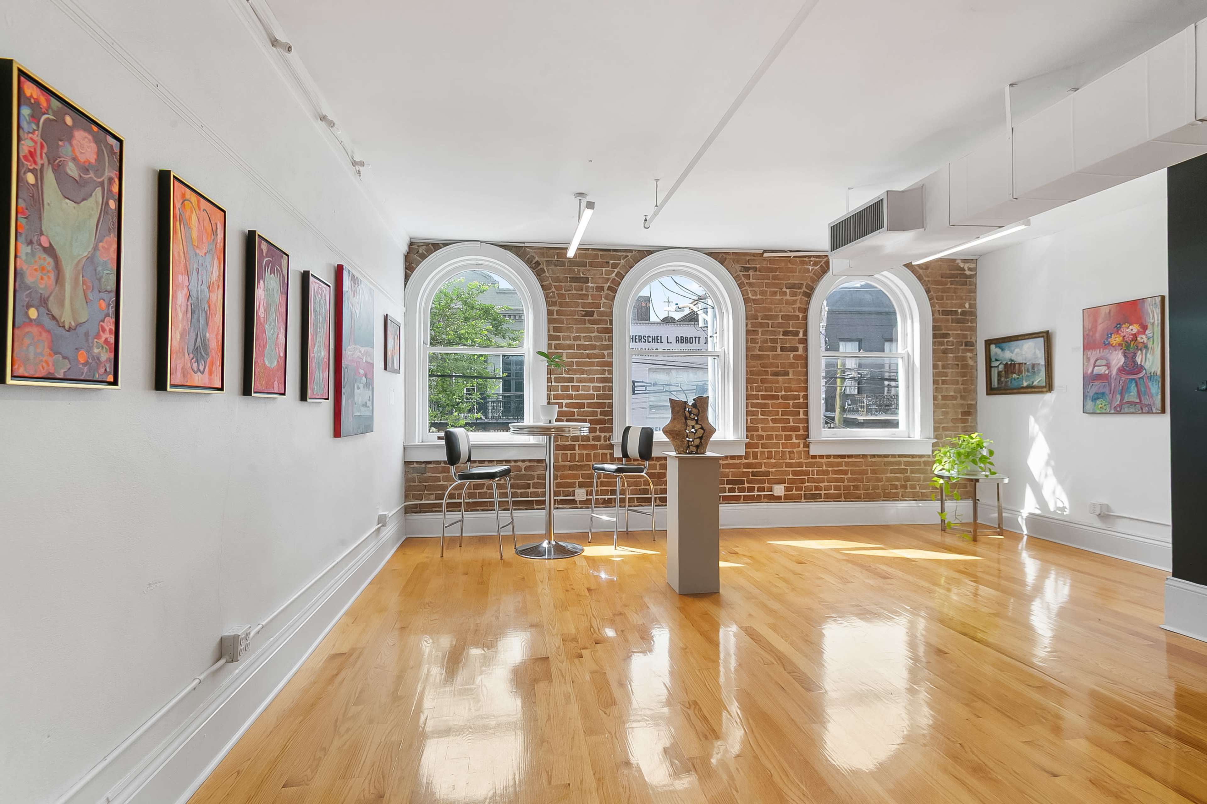 The image shows an art gallery with brick walls, featuring several framed artworks hung along one side and a small seating area with two stools.
