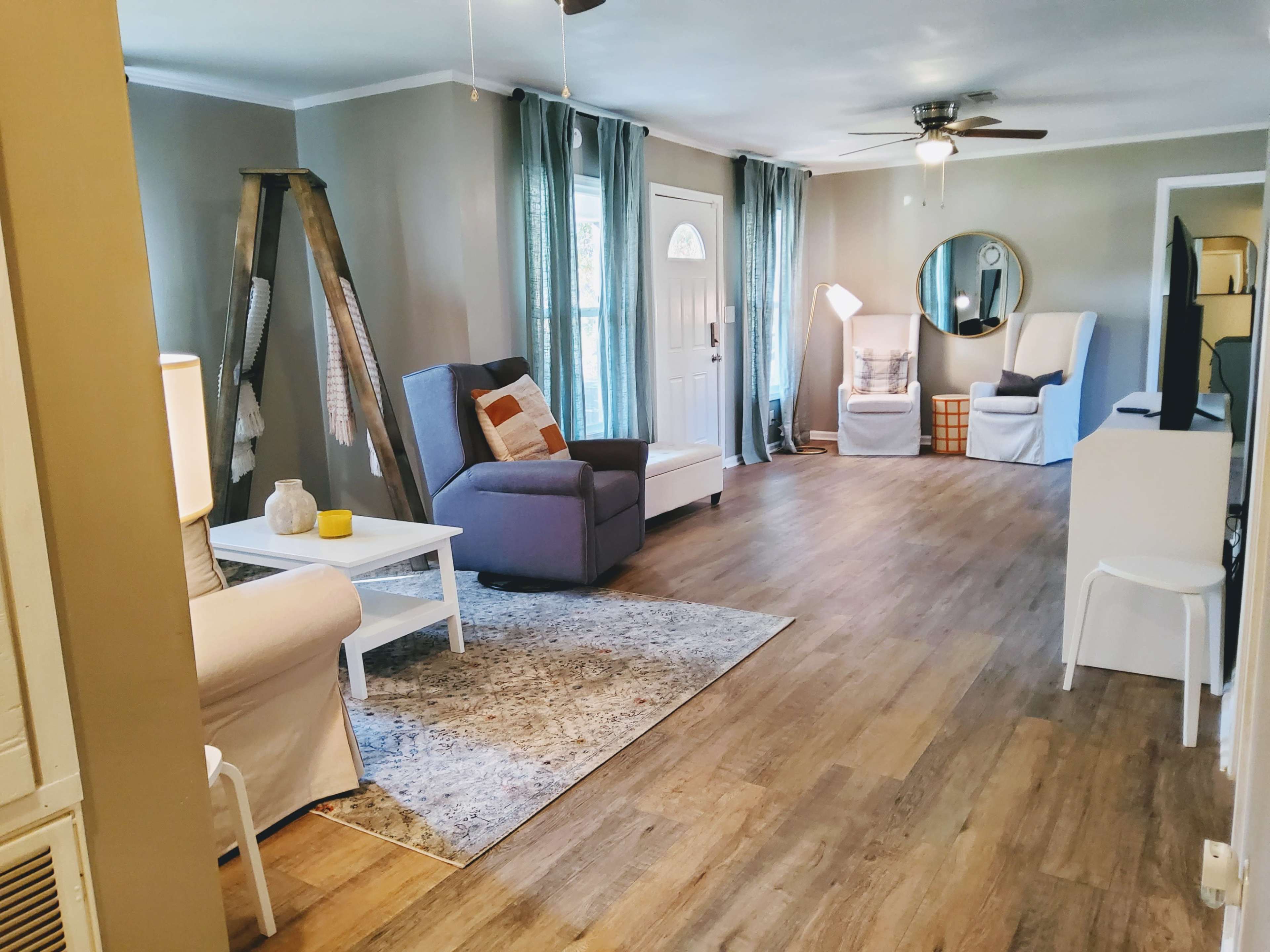 A spacious living room with wooden floors, a ceiling fan, two armchairs, a coffee table, and large windows covered by curtains.