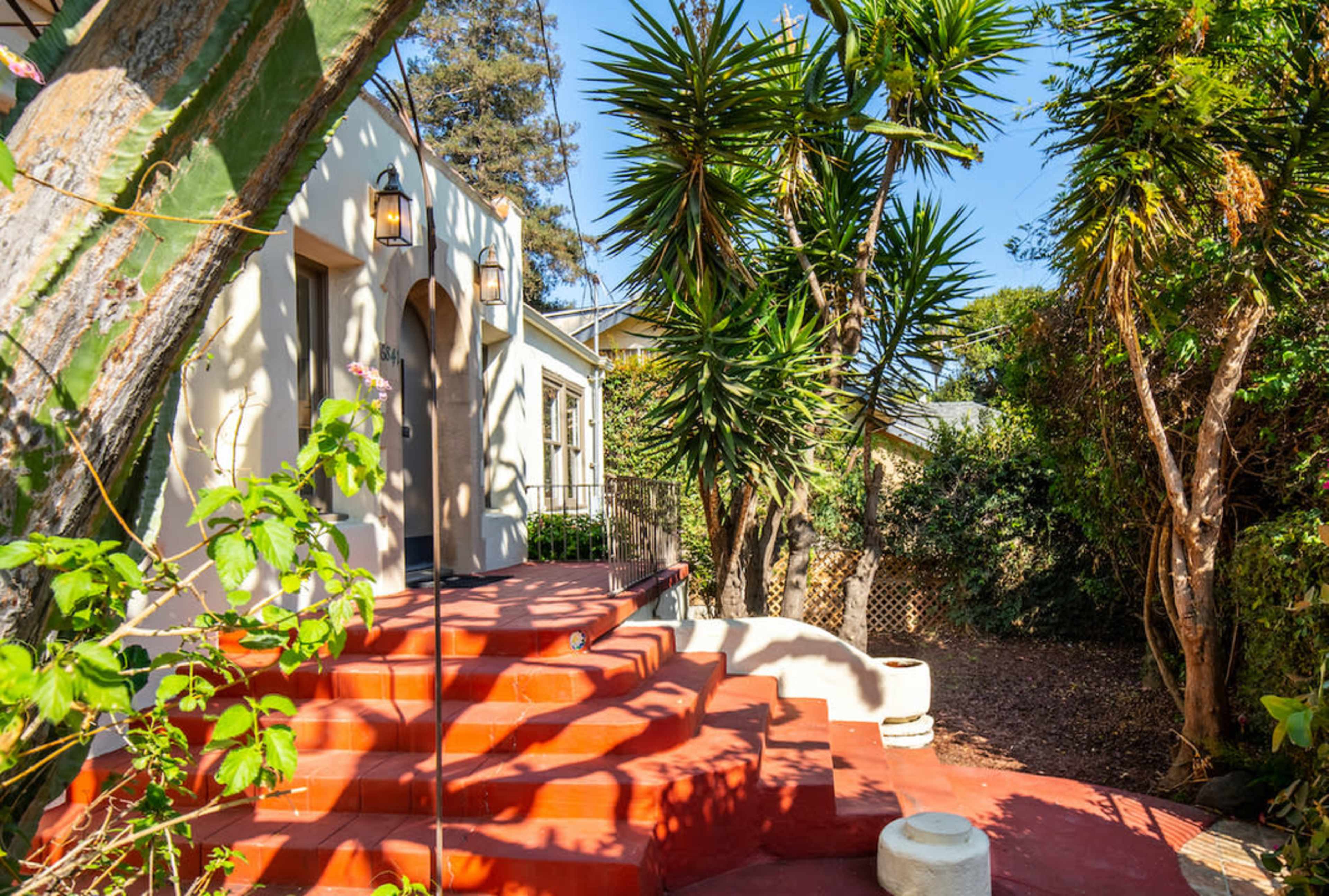 The image shows a house entrance with red stairs flanked by lush greenery and palm trees.
