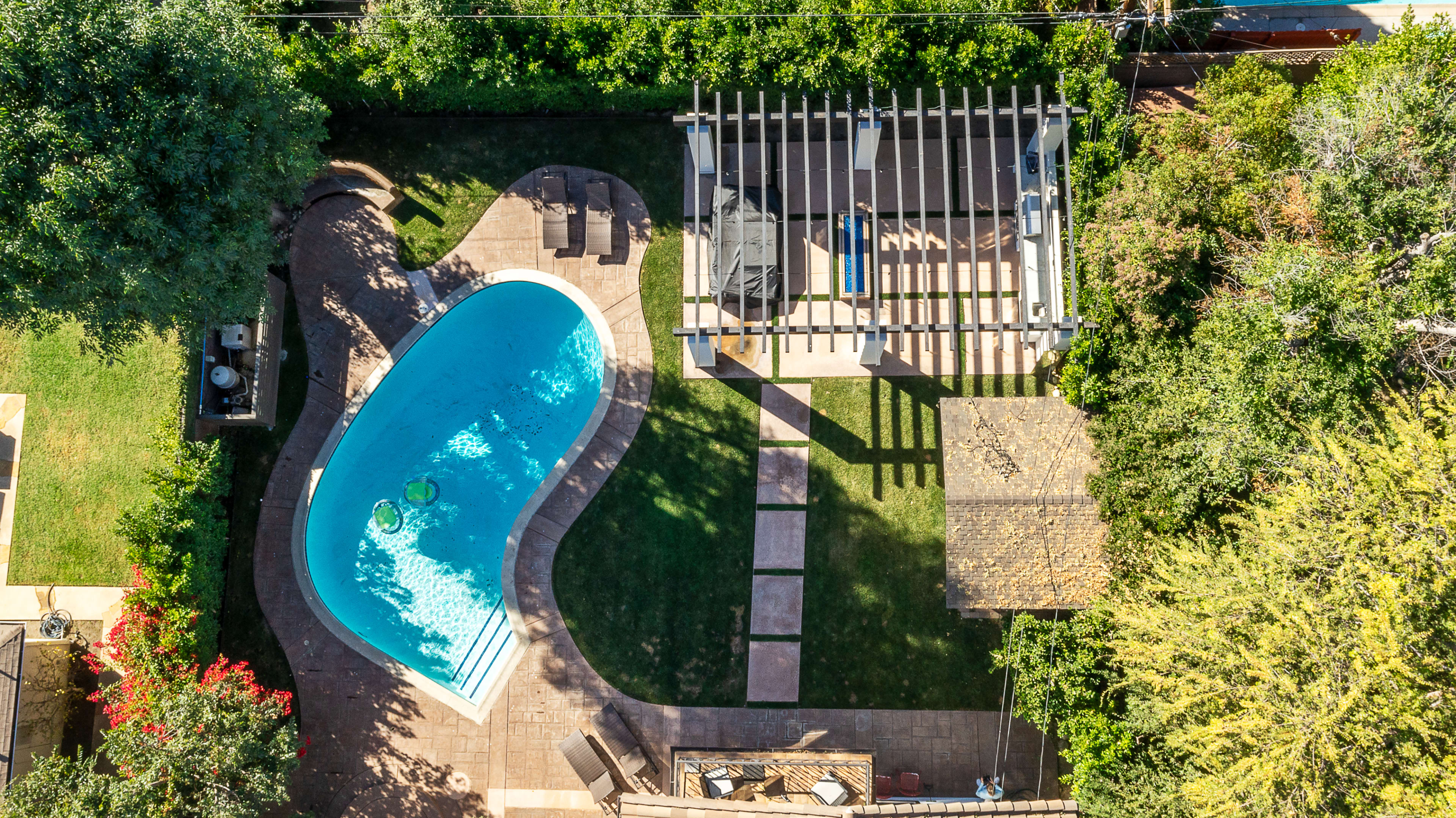 An aerial view of a backyard featuring a curved swimming pool, a covered patio area, and landscaped greenery.