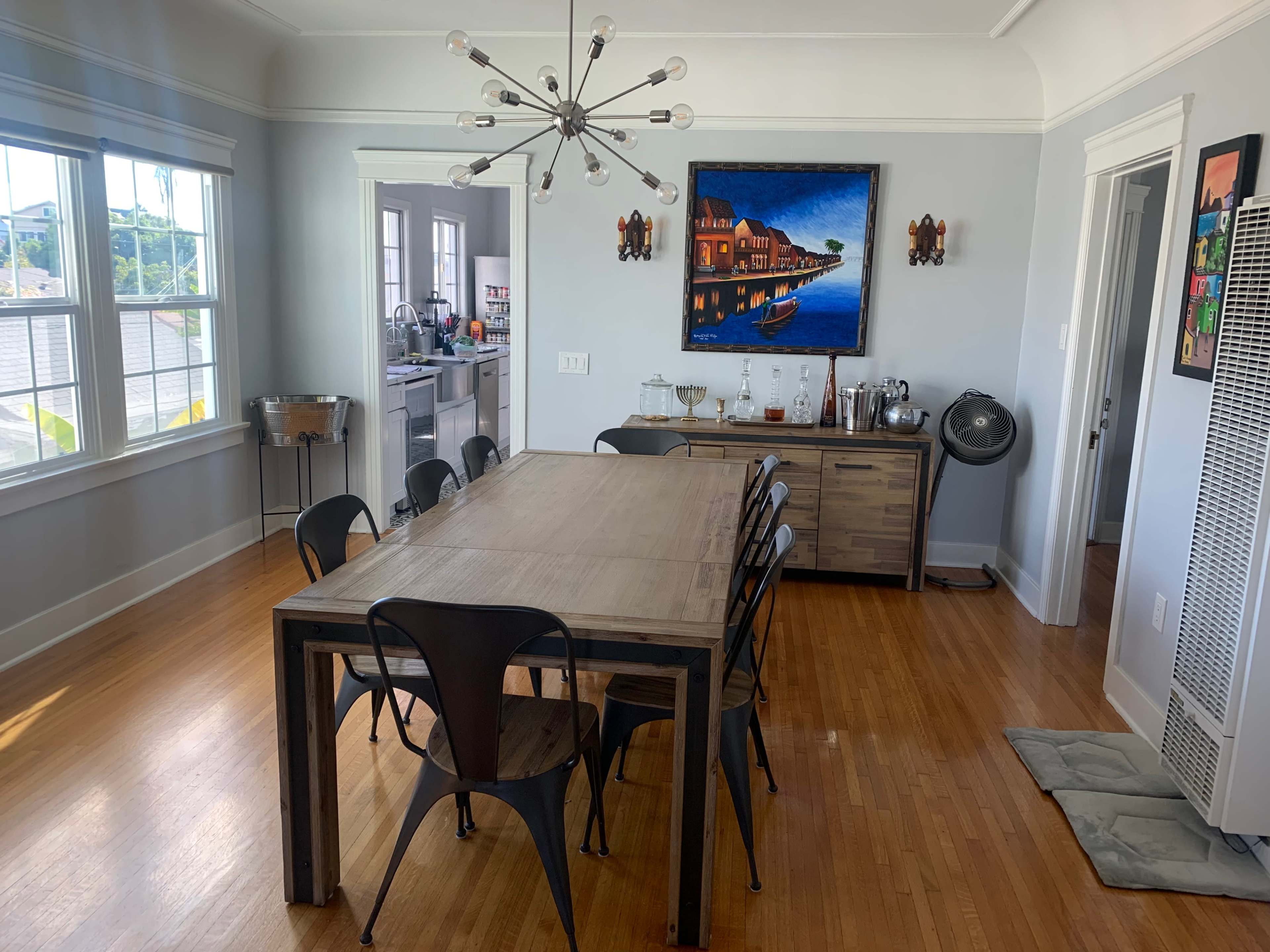 A modern dining room features a large wooden table surrounded by black chairs, with a colorful painting on the wall and a sideboard displaying glassware.
