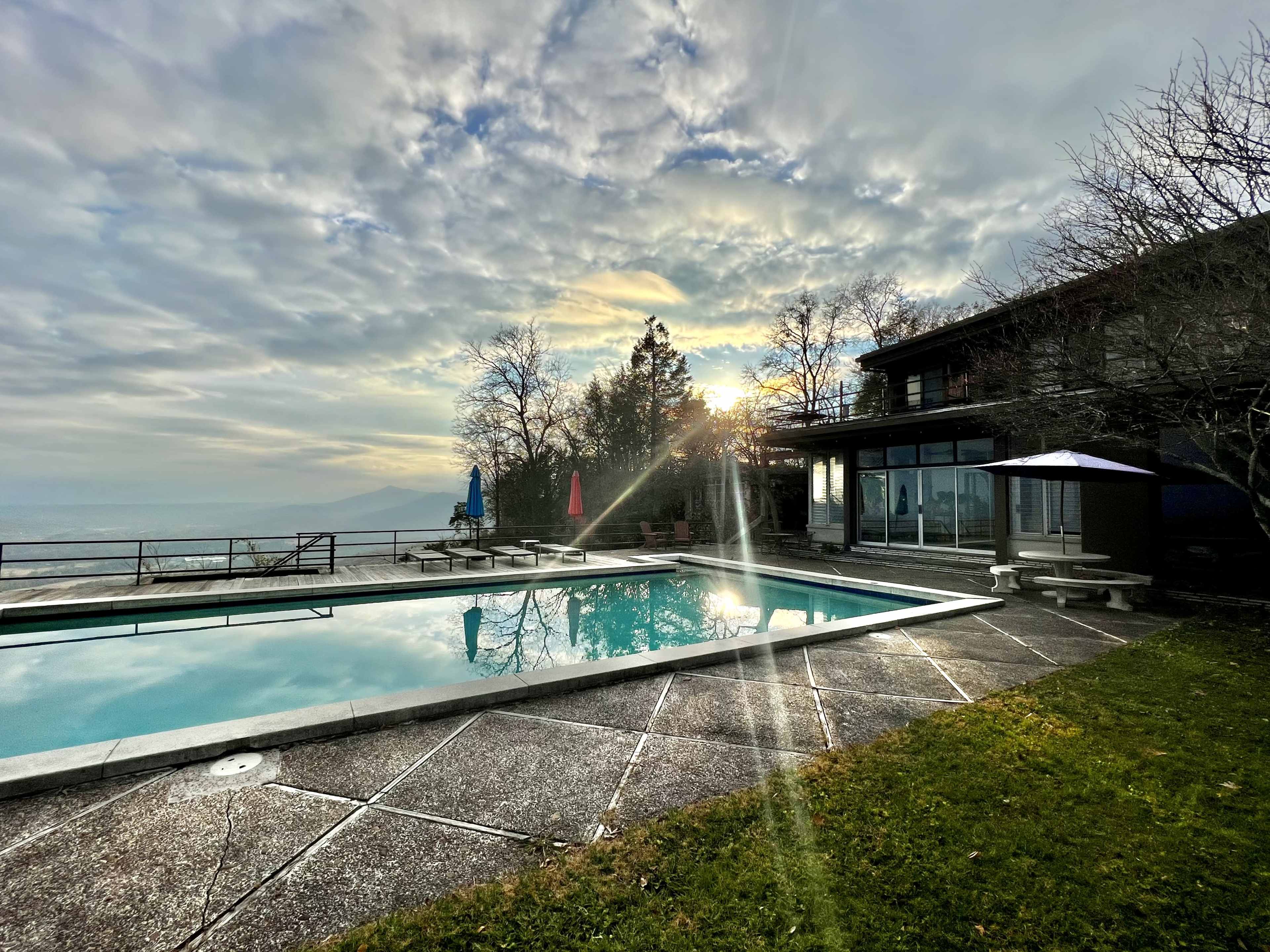 A rectangular swimming pool is situated beside a modern house with large windows, framed by trees and bathed in the light of a setting sun.