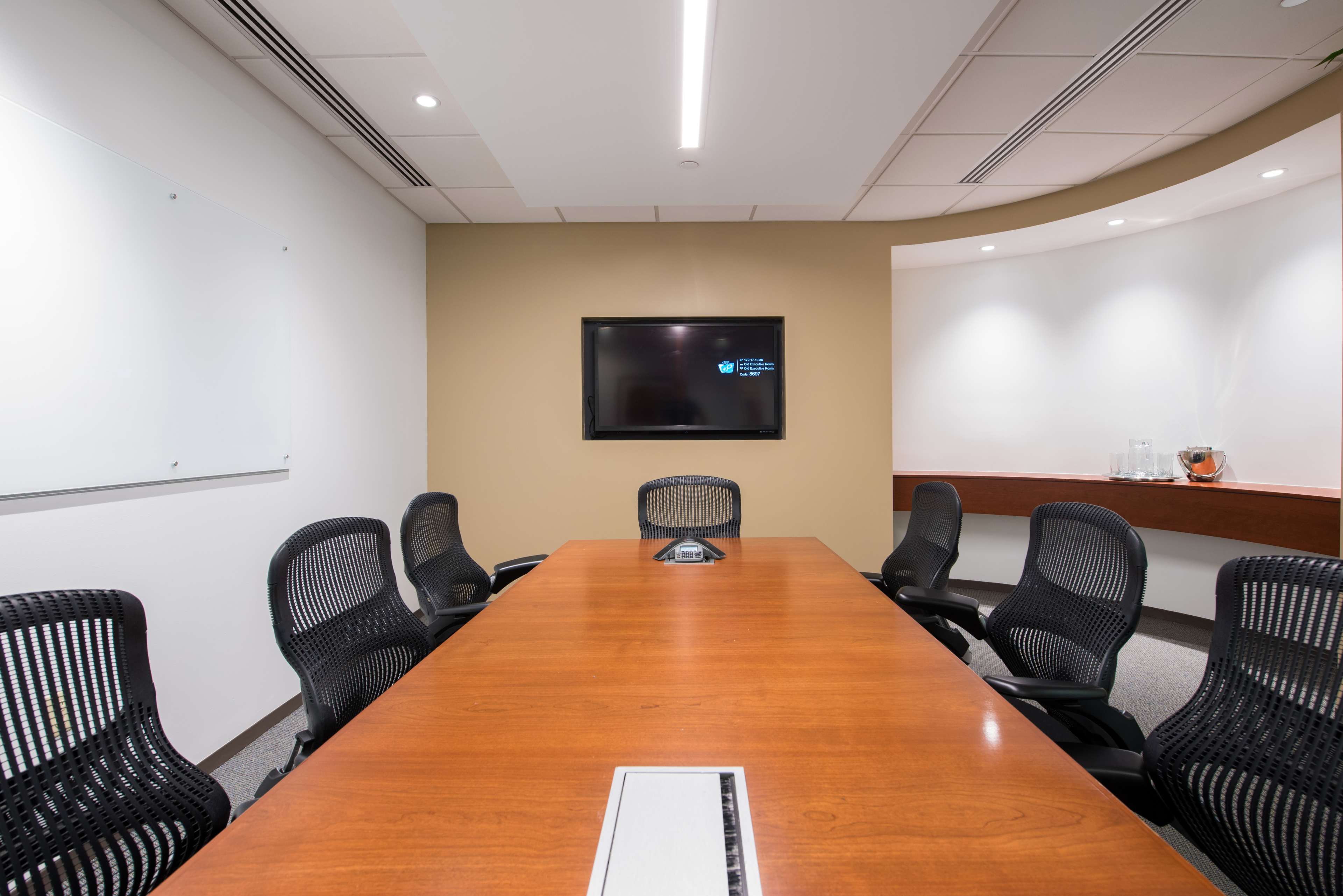 A conference room features a large wooden table surrounded by black ergonomic chairs, with a wall-mounted screen and a whiteboard.