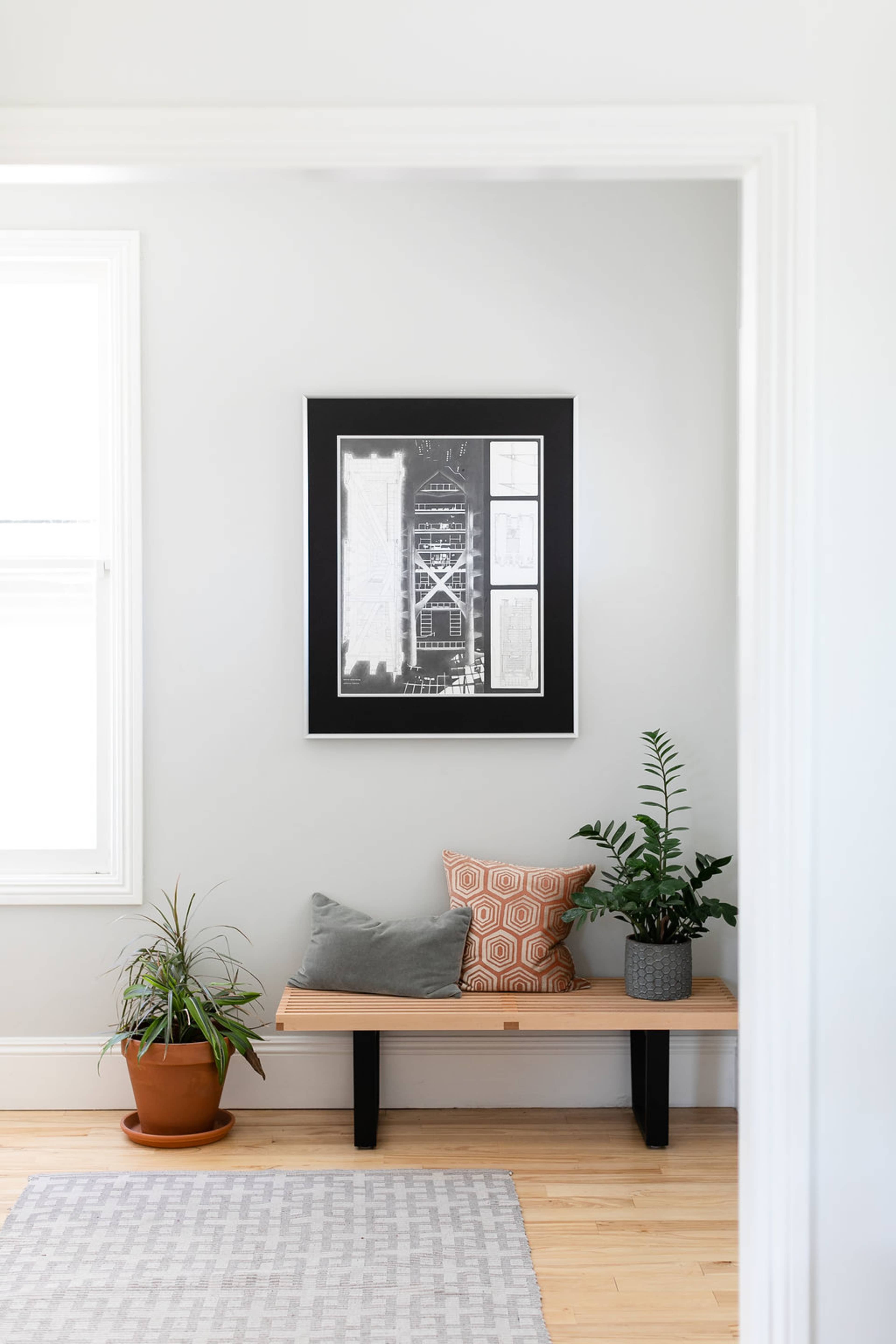 The image shows a minimalist interior space featuring a wooden bench with cushions, plants, and a framed artwork on the wall.
