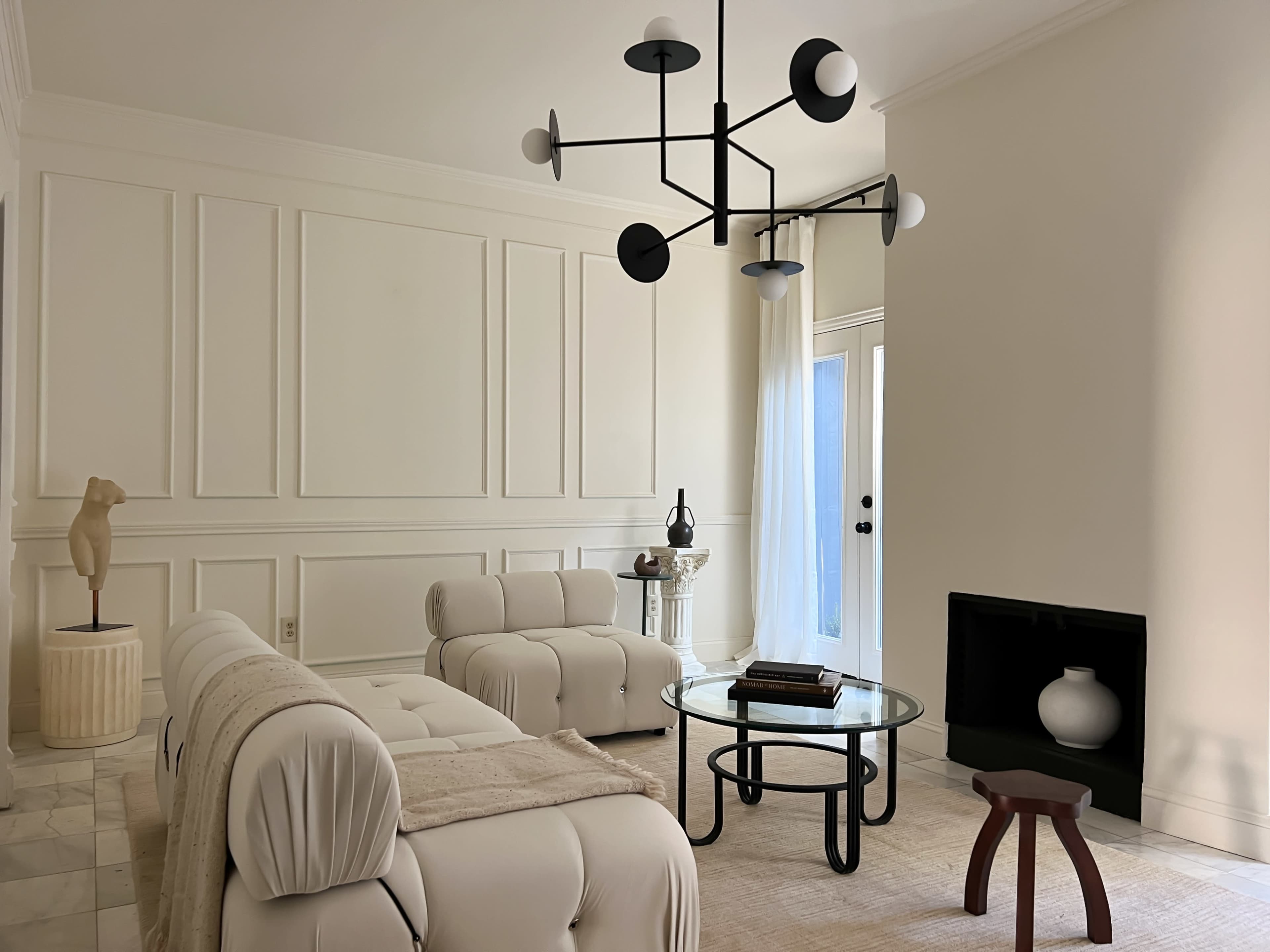 A minimalist living room features a white sectional sofa, a circular glass coffee table, and a modern chandelier, with a decorative sculpture and a black fireplace.