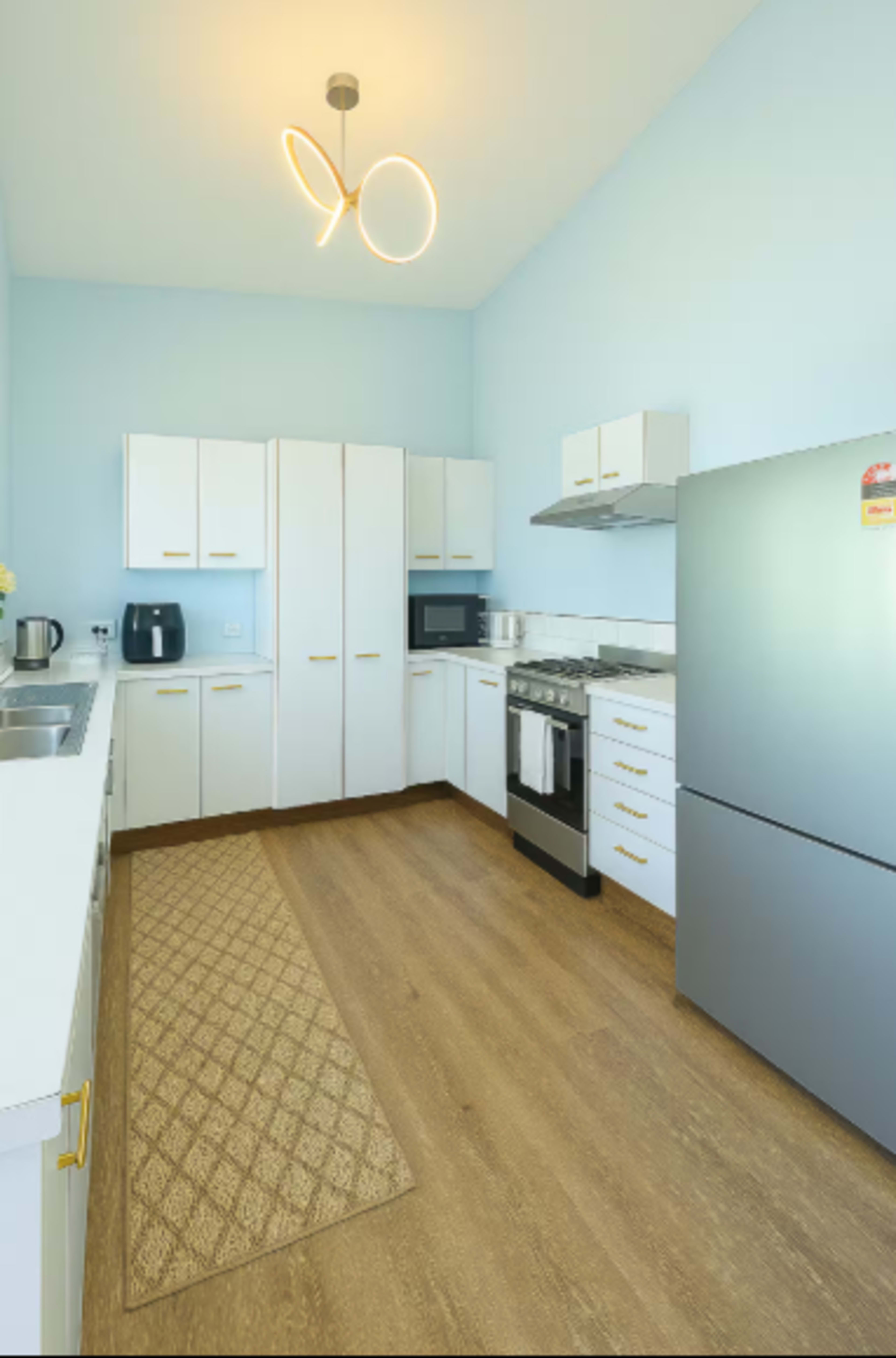 The image shows a modern kitchen with white cabinets, a gray refrigerator, a stove, and a patterned rug on a wooden floor.