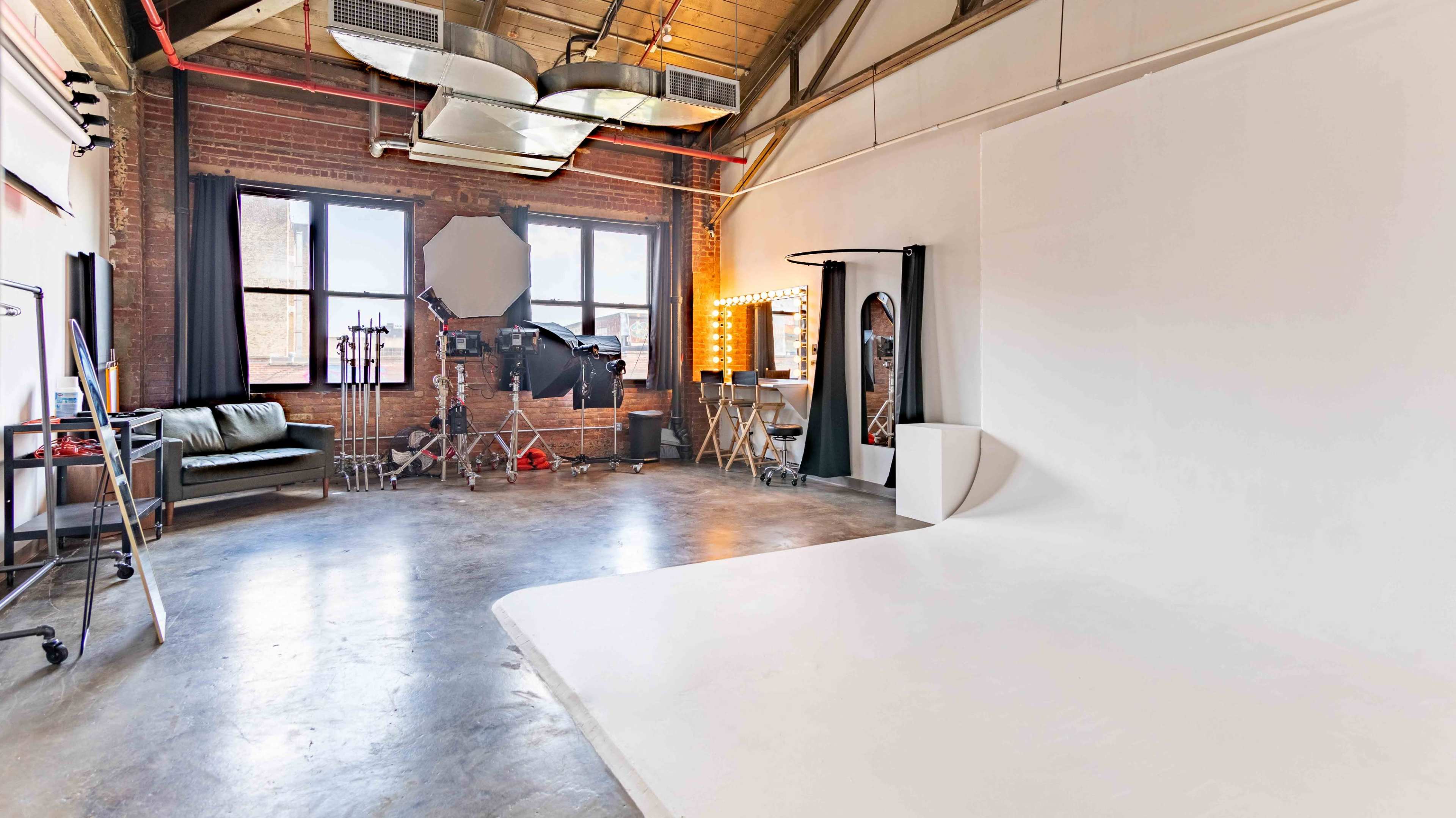 The image shows a spacious photography studio with brick walls, large windows, and a white seamless backdrop.