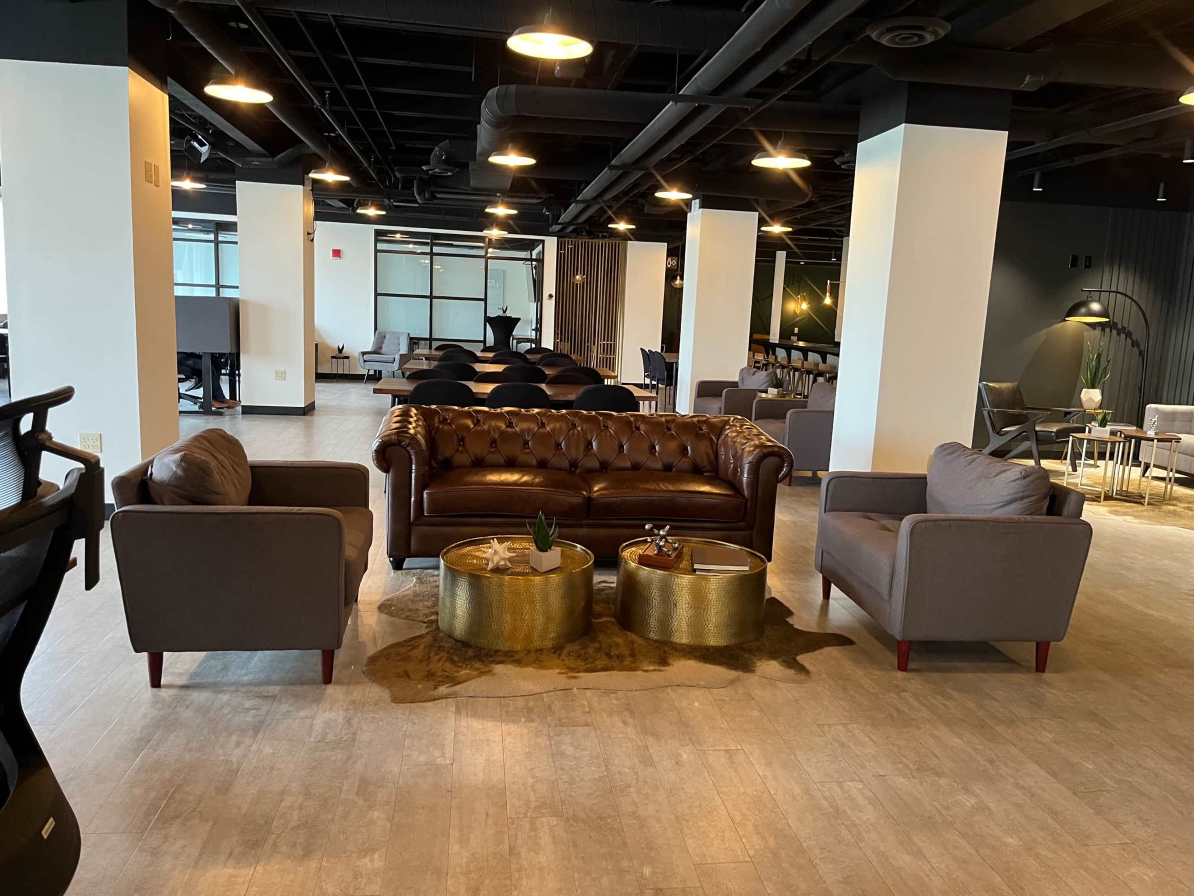 A modern lounge area featuring a brown leather sofa and two gray armchairs around gold coffee tables on a wooden floor.