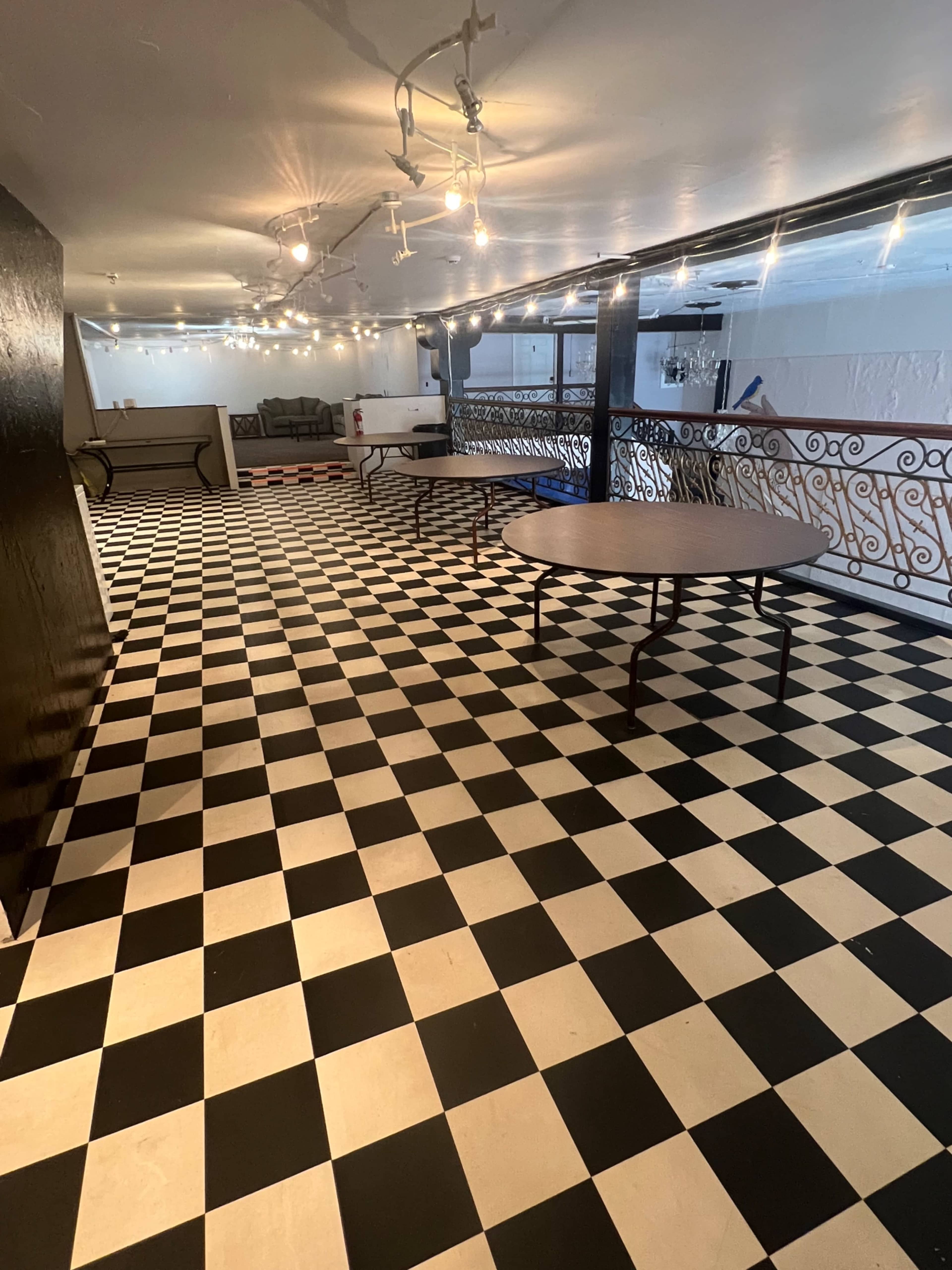The image shows a spacious room with a black and white checkered floor, tables arranged throughout, and string lights overhead.
