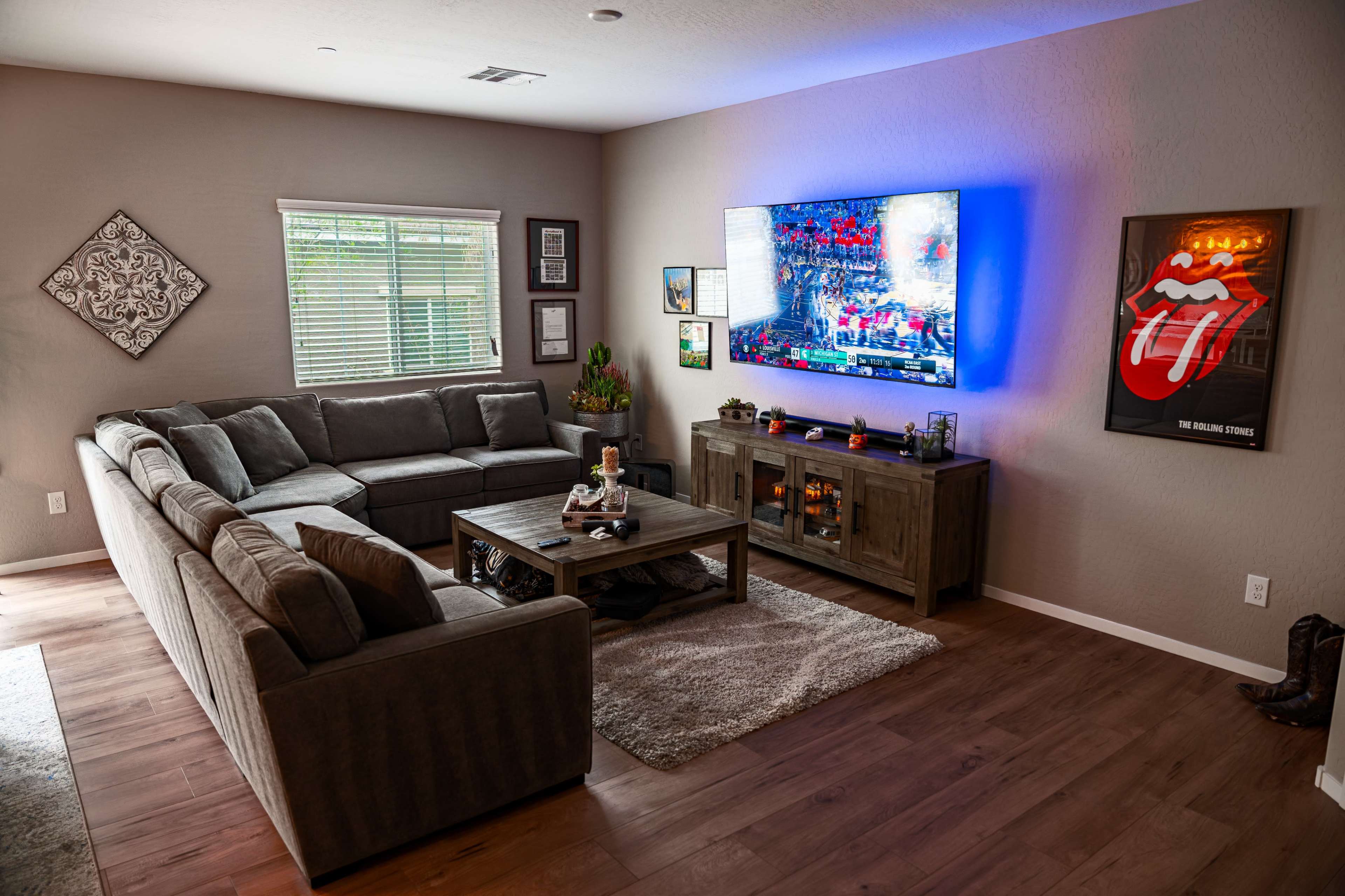 A cozy living room features a large sectional sofa, a wooden coffee table, and a mounted flat-screen TV displaying vibrant colors, with various wall decorations and a window allowing natural light.