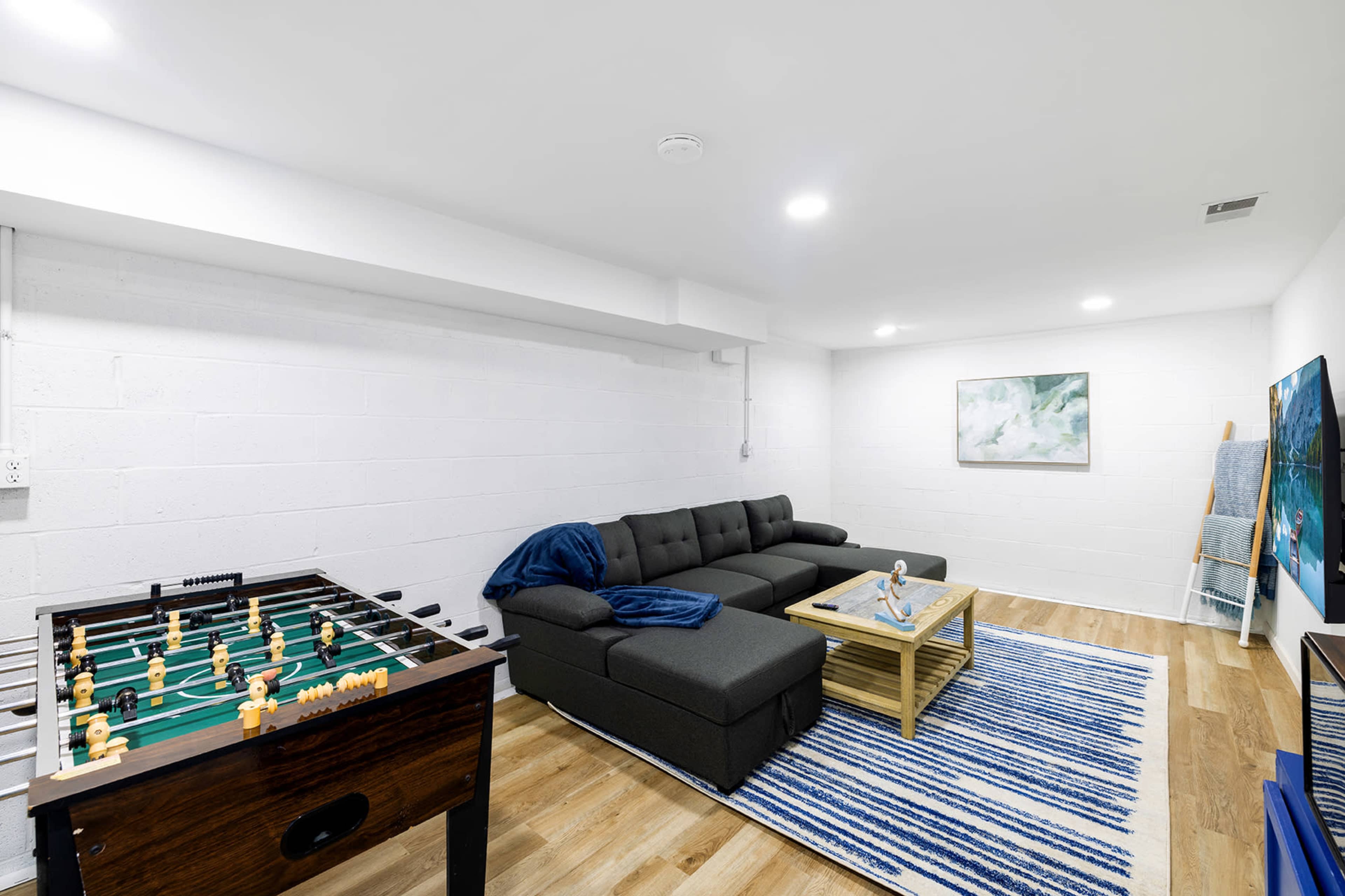 The image shows a modern basement room with a foosball table, a sectional sofa, a coffee table, and wall art.
