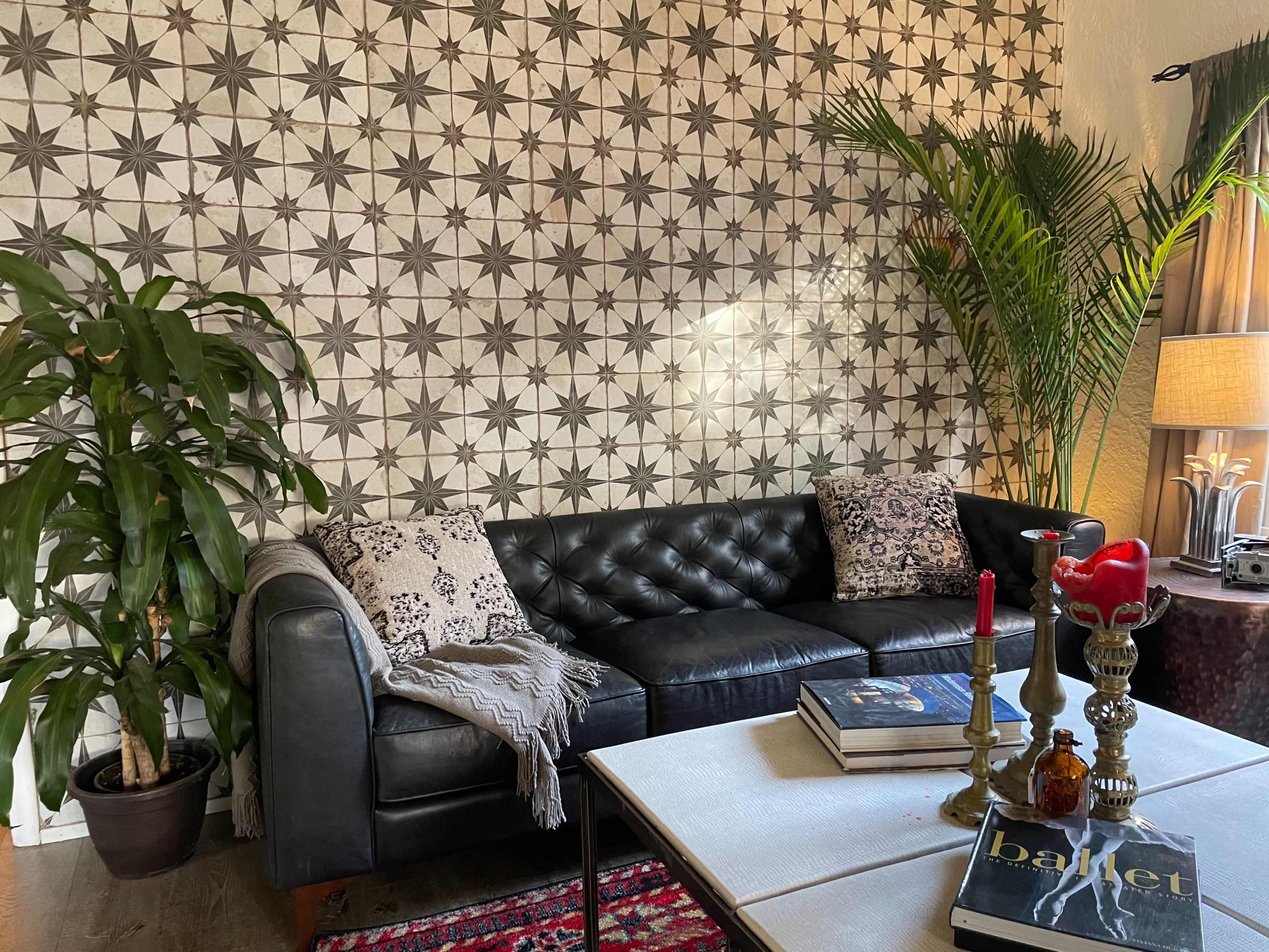 A black leather sofa is positioned in front of a patterned wall, accompanied by a plant, books, candles, and decorative pillows on a coffee table.