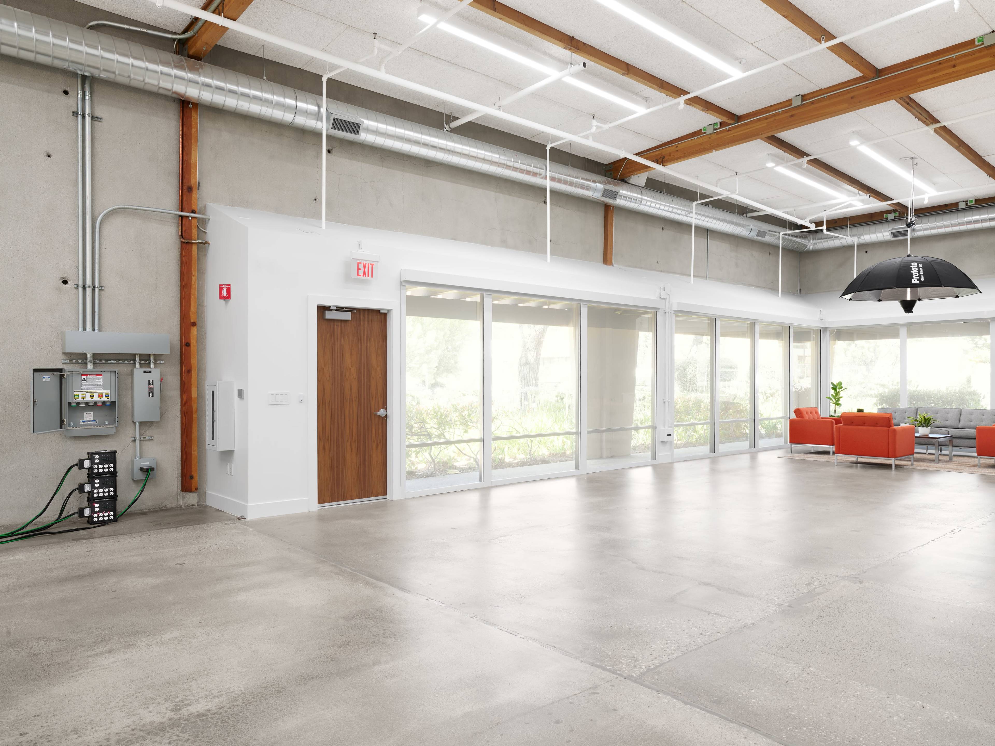 The image shows a spacious, modern interior with concrete flooring, large windows, and orange seating arrangements.