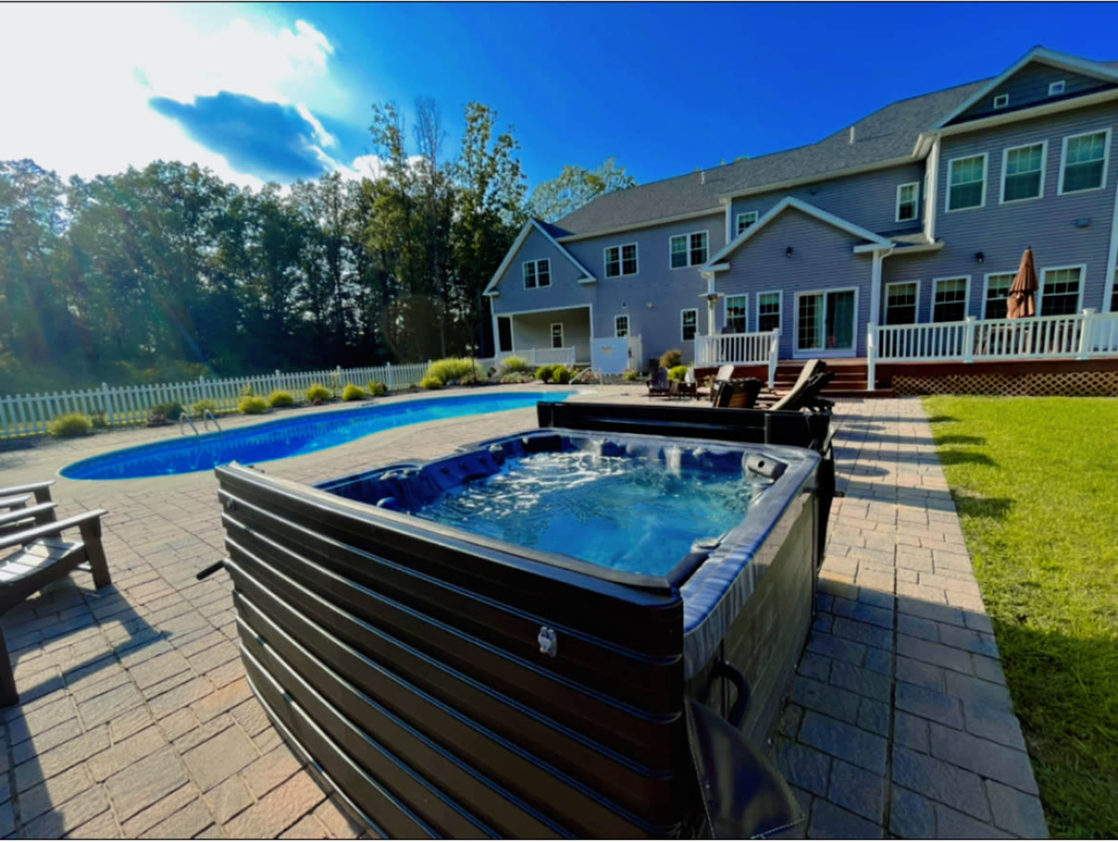 A backyard with a hot tub and a blue swimming pool surrounded by a paved patio and a large house in the background.