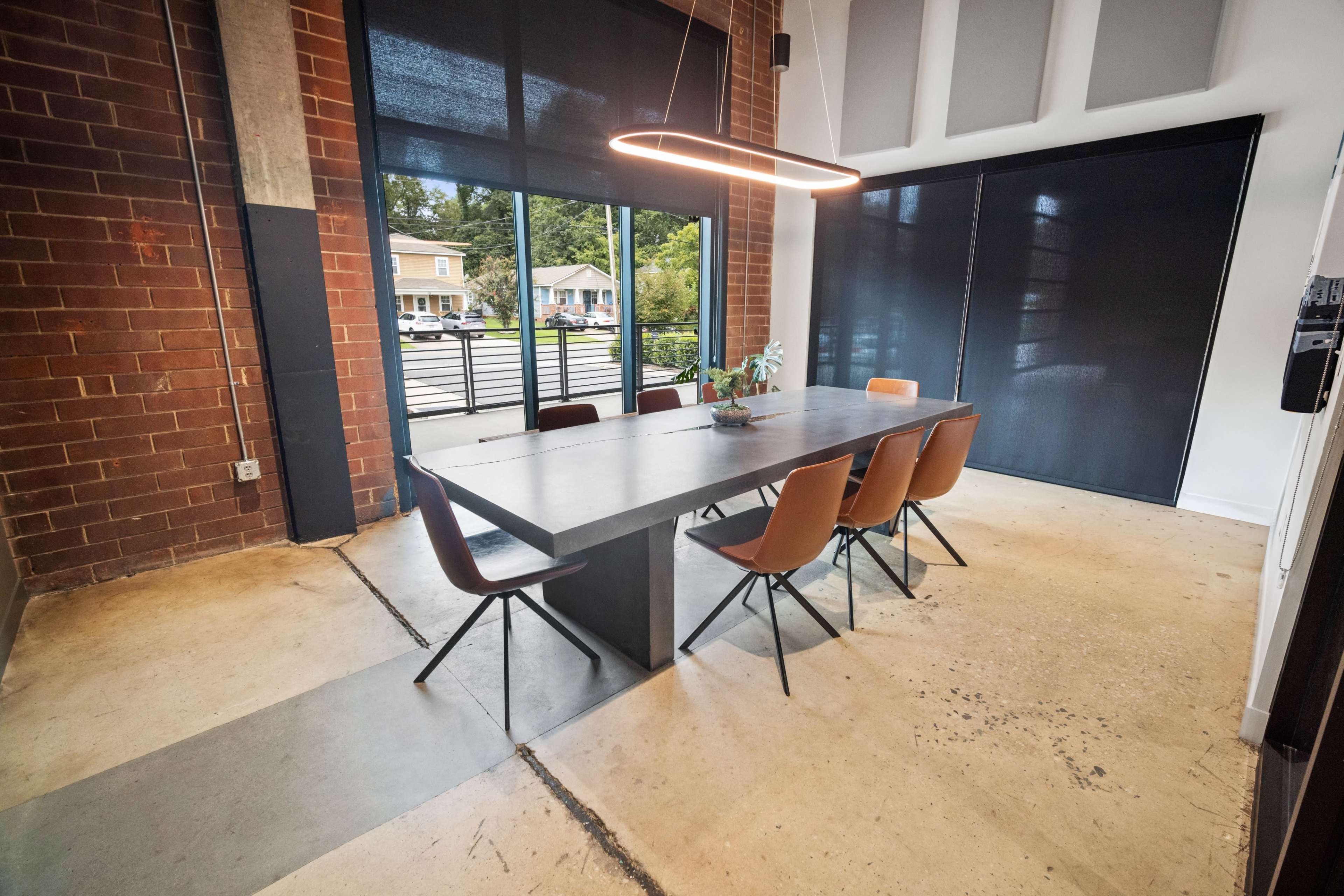 The image shows a modern dining area with a rectangular table and five chairs, set against a brick wall with large windows covered by black shades.