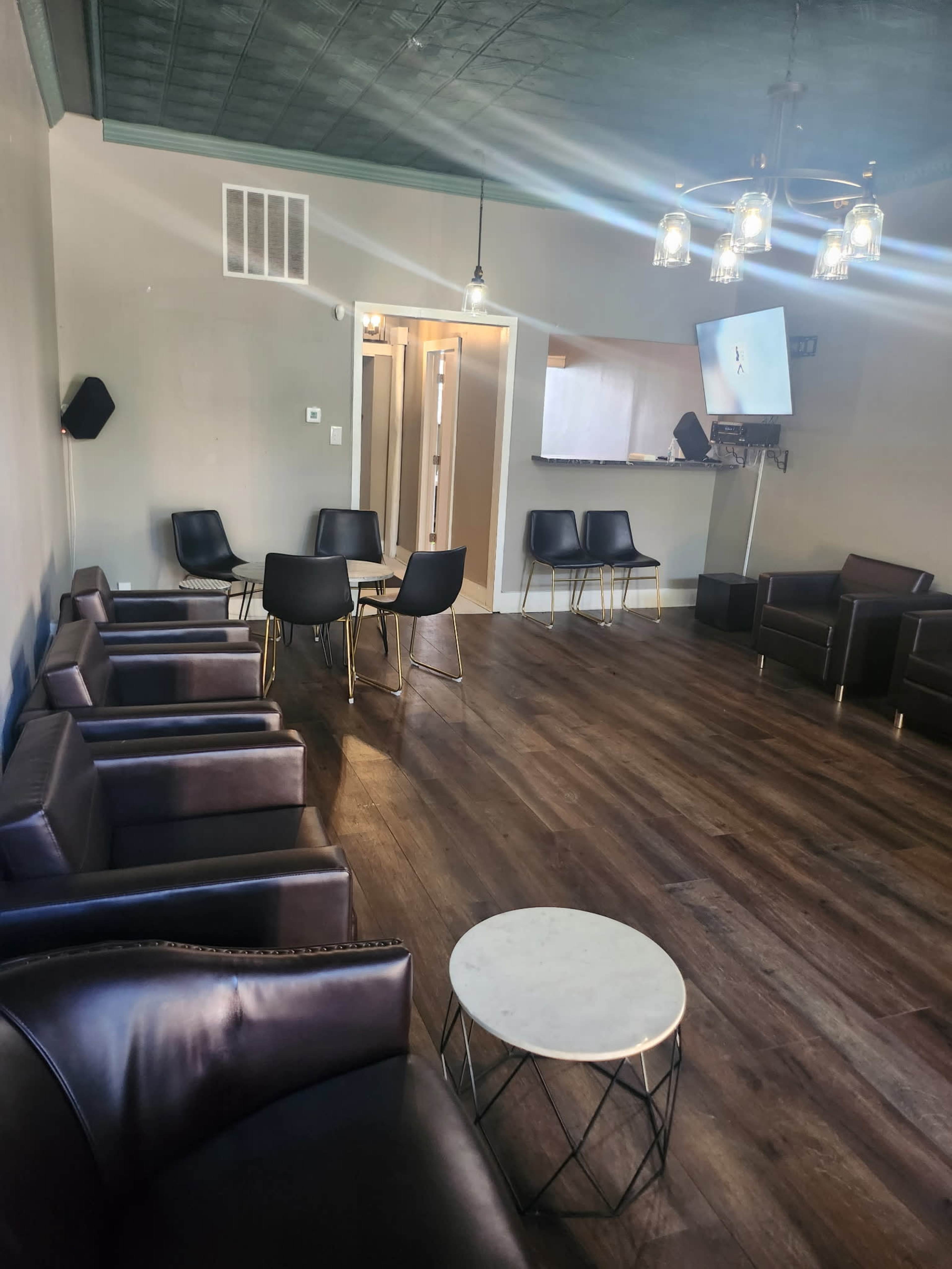 The image shows a modern lounge area with black chairs, a round white table, and wood flooring, featuring a television mounted on the wall.