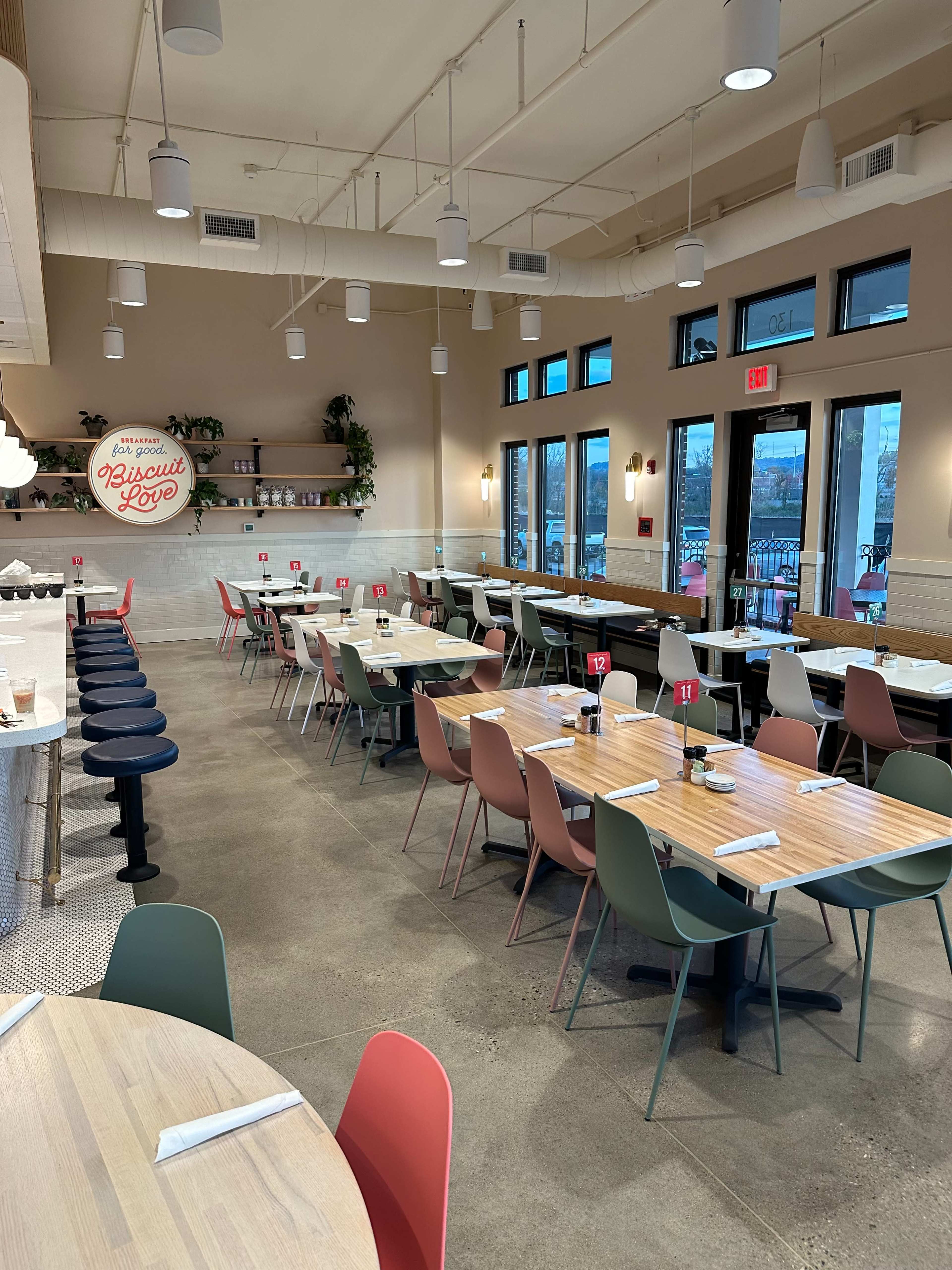 The image shows a modern restaurant interior with a mix of wooden tables and colorful chairs arranged throughout the dining area, featuring large windows that let in natural light.